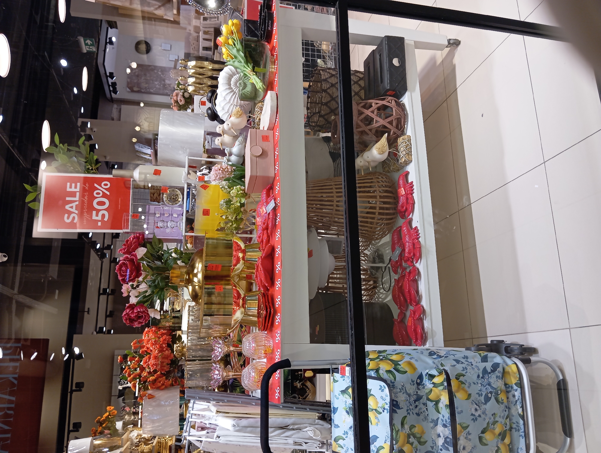 This is a photograph of a store window display in Wołomin, Poland. The display features a white table laden with various decorative items. On the table, there are several red lobster-shaped decorations, a pink jewelry box, white ceramic birds, a white seashell-shaped object, a glass vase with yellow tulips, and a black tissue box with a "Meow" label. A red and white patterned runner with "%" symbols covers the front of the table. Behind the table, there are more decorative items, including gold vases and pink and red flowers. A red "SALE -50%" sign is visible in the upper right portion of the display. To the left of the table, a rolling shopping cart with a blue and yellow lemon-patterned bag is positioned. The floor is tiled in a light beige color. Reflections of lights and interior store elements are visible in the glass.