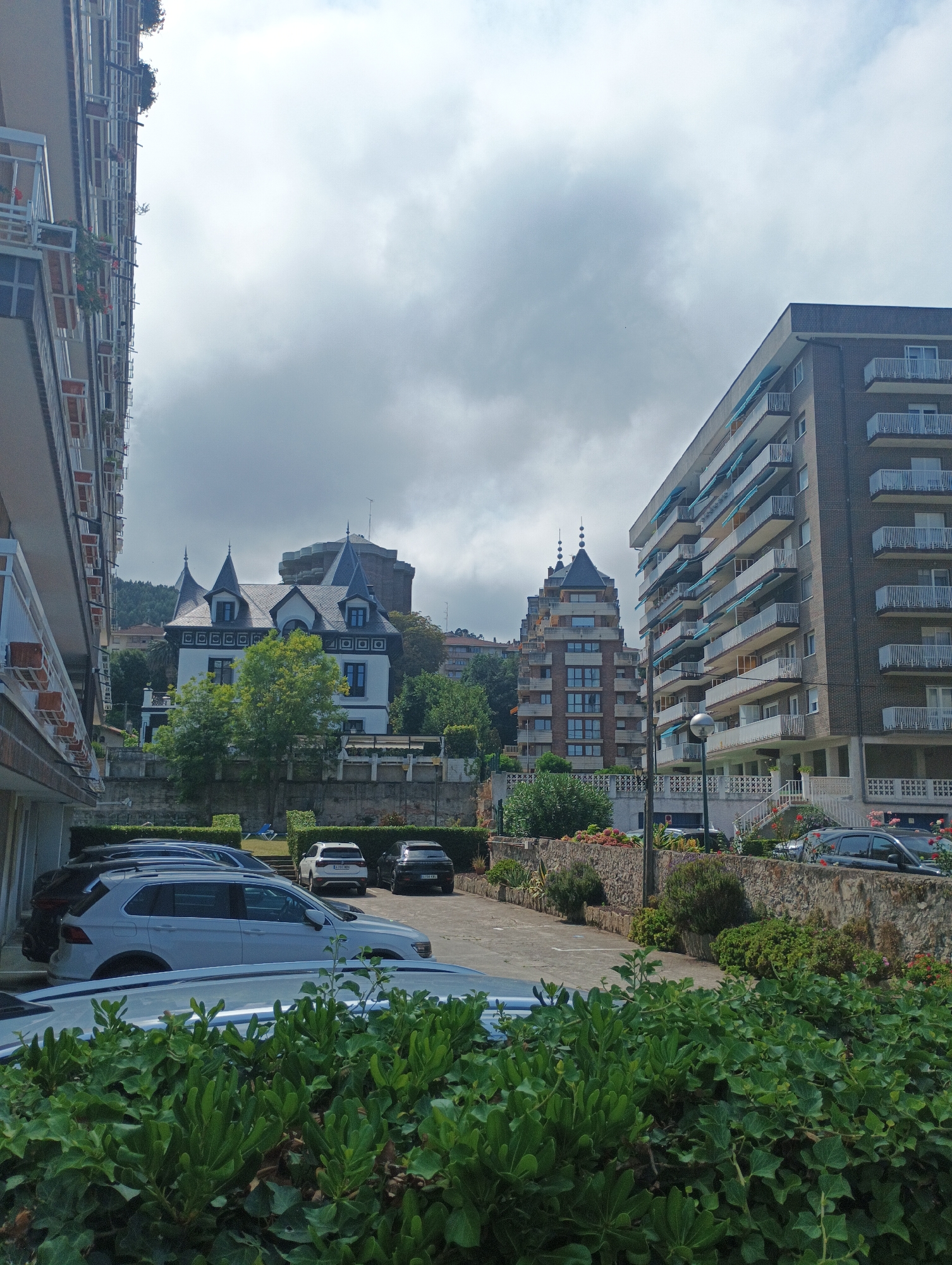The image depicts a view of buildings and parked cars in Castro-Urdiales, Spain. In the foreground, a dense green bush partially obscures the view of several parked cars, including a white SUV. To the left, the side of a modern apartment building with balconies is visible, extending upwards out of frame. In the mid-ground, several other buildings are present. A distinct white villa-style house with dark roofs and ornate trim is situated on a hill, flanked by trees. Behind it, taller, more modern apartment buildings with many balconies can be seen. The sky is overcast with thick, grey clouds. The scene appears to be a residential area with a mix of architectural styles.