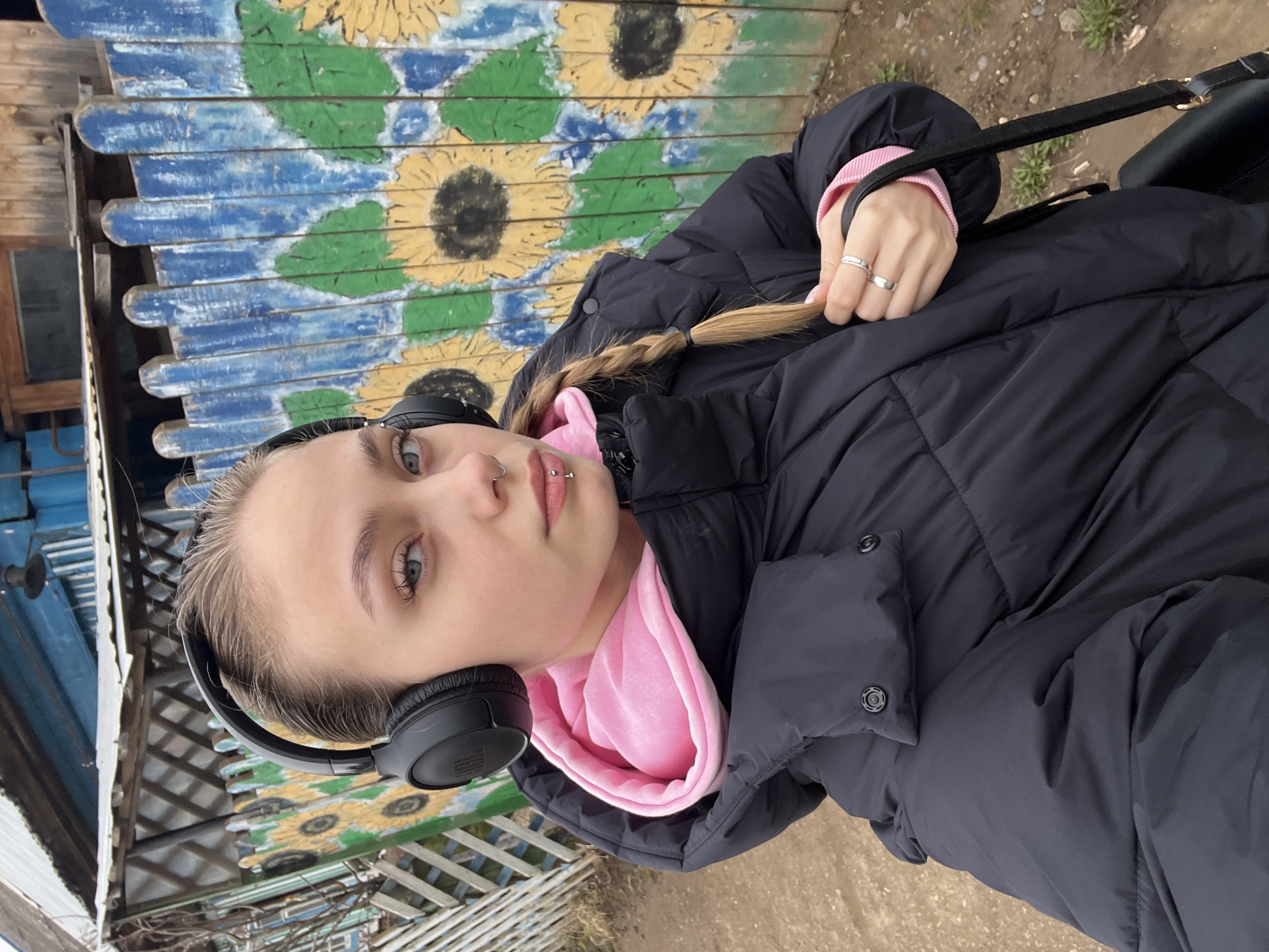 A young woman stands outdoors, wearing a black puffer jacket over a pink hooded sweatshirt, and black headphones. She has fair skin, light eyes, and blonde hair styled in a braid, which she is holding with her right hand, adorned with two rings. Her facial features include pierced nostrils and a double lip piercing.
Behind her is a wooden structure decorated with painted sunflowers in shades of yellow, green, and blue. The background suggests an urban environment, possibly in Фоки, Russia, given the context. The ground appears to be a mix of grass and dirt. She's also carrying a dark-colored bag over her shoulder.