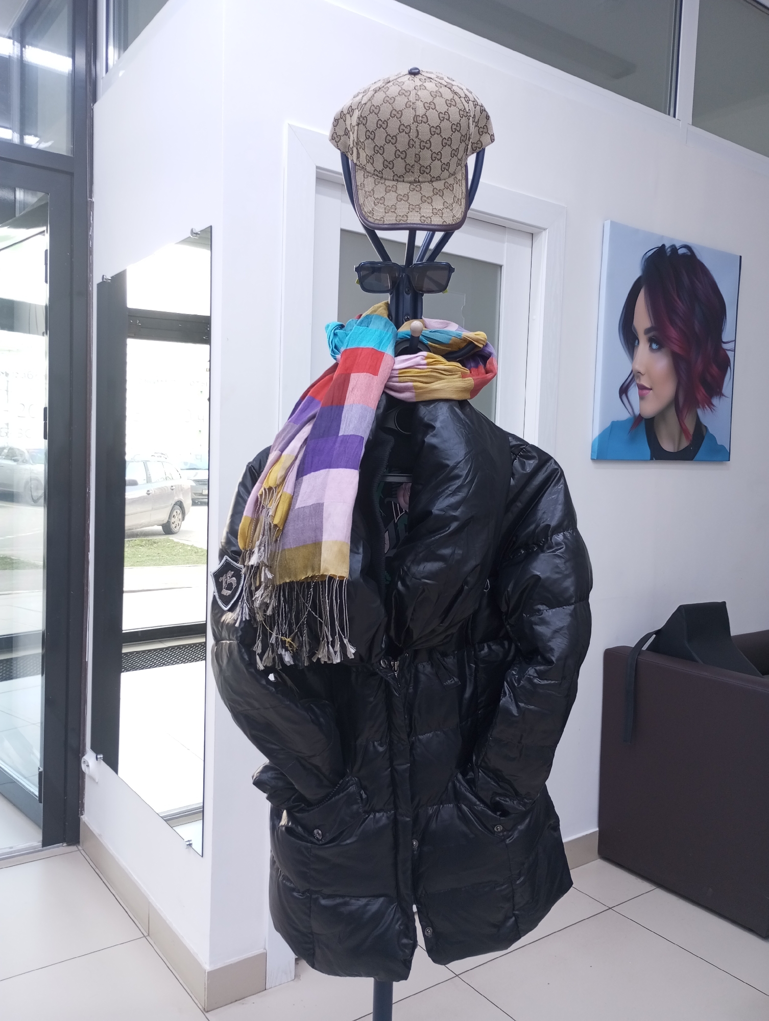 This media file captures an indoor scene, likely an entryway or waiting area, in Izhevsk, Russia. The central focus is a black, glossy puffer coat hanging on a dark metal coat rack. Perched atop the coat rack is a beige baseball cap adorned with a repeating "GG" pattern, indicating a Gucci brand. Below the cap, a pair of dark sunglasses hangs, followed by a vibrant, multi-colored checkered scarf featuring shades of purple, blue, green, yellow, orange, and red, complete with tassels.
To the left of the coat rack, a tall glass door or window provides a view of the exterior, reflecting a light-colored car, possibly a silver or beige wagon, parked outdoors. A small, rectangular mirror is mounted on the white wall next to the door.
On the white wall to the right, a framed artwork or poster depicts the profile of a woman with striking blue eyes and short, wavy red hair, looking towards the left. Below this artwork, a section of a dark brown armchair or sofa is visible, suggesting a seating area.
The scene is brightly lit, indicative of daytime. The presence of a puffer coat and scarf suggests cooler weather or an indoor environment where outerwear is stored. The overall arrangement implies a commercial establishment, possibly a salon or beauty parlor, given the artwork of the styled woman.