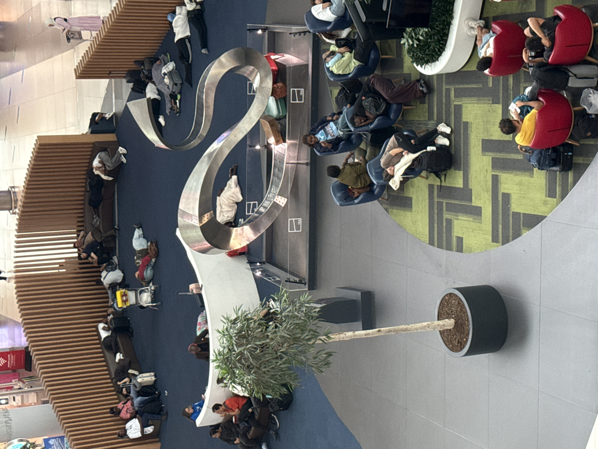 This is an aerial, high-angle, full-shot photograph of an airport terminal in Arnavutköy, Turkey, likely at Istanbul Airport, given the overall aesthetic and scale. The scene captures a large, open-plan waiting area with various seating arrangements and architectural elements. The floor is a mix of dark blue carpet and grey tiles.
In the background, a curved wooden slatted wall divides the space. Along this wall, several people are resting or sleeping on a long, dark-colored sofa. Luggage, including suitcases and bags, is scattered around them. A yellow luggage cart is also present. Further back, near a beige wall, a person in a pink dress is visible standing near a luggage cart, suggesting an active traveler amidst the resting individuals.
In the mid-ground, a large, metallic, ribbon-like sculpture dominates the center of the space. Below and around this sculpture, numerous people are lying down and sleeping directly on the blue carpet, some with blankets or sleeping on their luggage. Various suitcases and bags are placed around them.
To the right of the sculpture, a curved white wall encloses a plant, and beyond that, more people are seated in blue armchairs, many of whom appear to be resting or sleeping. Some have their heads leaning back or on their arms, and luggage is at their feet.
In the foreground, a potted tree with a slender trunk and leafy branches stands in a dark cylindrical planter on the grey tiled floor. To the right of the tree, a green and black patterned carpet section is visible. On this carpet, several red, egg-shaped chairs are occupied by people, some of whom are reading or resting. Luggage is visible around these chairs as well. A black television screen is mounted on a stand to the right.
The overall impression is one of a busy airport environment where travelers are waiting, with many choosing to sleep due to delays or long layovers. The lighting suggests it is daytime, with ambient indoor lighting illuminating the space.