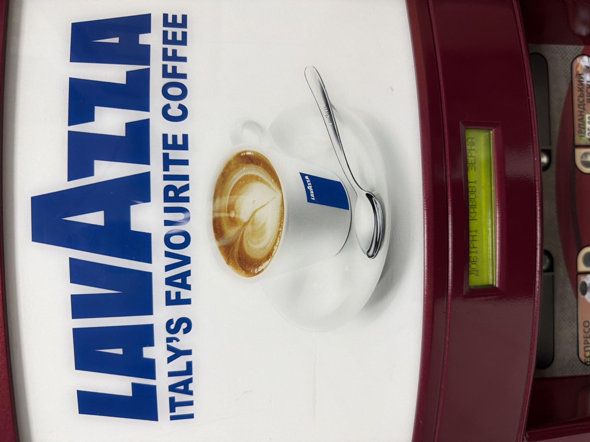 A close-up, rotated shot captures the front panel of what appears to be a Lavazza coffee vending machine. The dominant feature is a large white panel on the left, adorned with prominent blue text. The word "LAVAZZA" is displayed in large, bold capital letters, with "ITALY'S FAVOURITE COFFEE" written in smaller blue capital letters beneath it, all oriented sideways due to the rotation of the image.
To the right of this text, a white ceramic coffee cup, complete with a handle, is shown resting on a saucer. The cup contains coffee with intricate latte art on its surface. A small blue label with "Lavazza" written in white is visible on the side of the cup. A silver spoon, engraved with "Lavazza" on its handle, lies on the saucer next to the cup.
The right side of the image reveals the dark red frame of the vending machine. Embedded within this frame is a rectangular, green digital display. The display shows text in Cyrillic script: "КАВОВІ ЗЕРНА", which translates to "COFFEE BEANS". Further to the right, partially visible text in Cyrillic indicates menu options like "ІРЛАНДСЬКИЙ" (Irish) and "ЕСПРЕСО" (Espresso), suggesting the variety of beverages offered by the machine. No people are visible, and the setting appears to be indoors. The image's rotation makes all elements appear sideways.