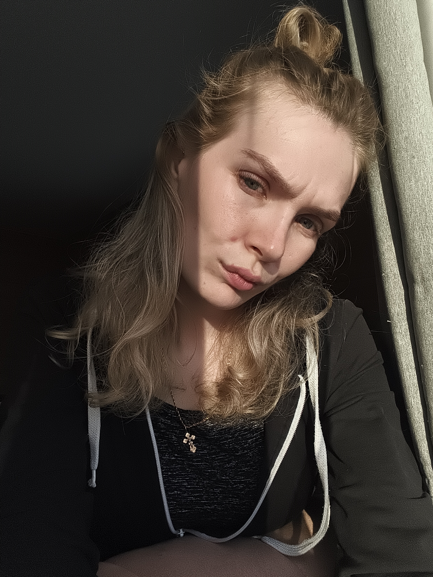 A blonde woman with fair skin is shown in a selfie. Her hair is partially up in a bun, and the rest is loose around her shoulders. She wears a dark-colored hooded sweatshirt over a patterned top and a delicate necklace with a cross pendant. She has a slight frown.
The background is ambiguous. To the left, there is darkness, possibly a wall or shadowed area. To the right, there is a textured, light-colored surface, possibly a curtain or wall. Strong, direct sunlight is illuminating one side of her face. The location is not discernible from the image and cannot be identified as Русский Вожой, Russia.