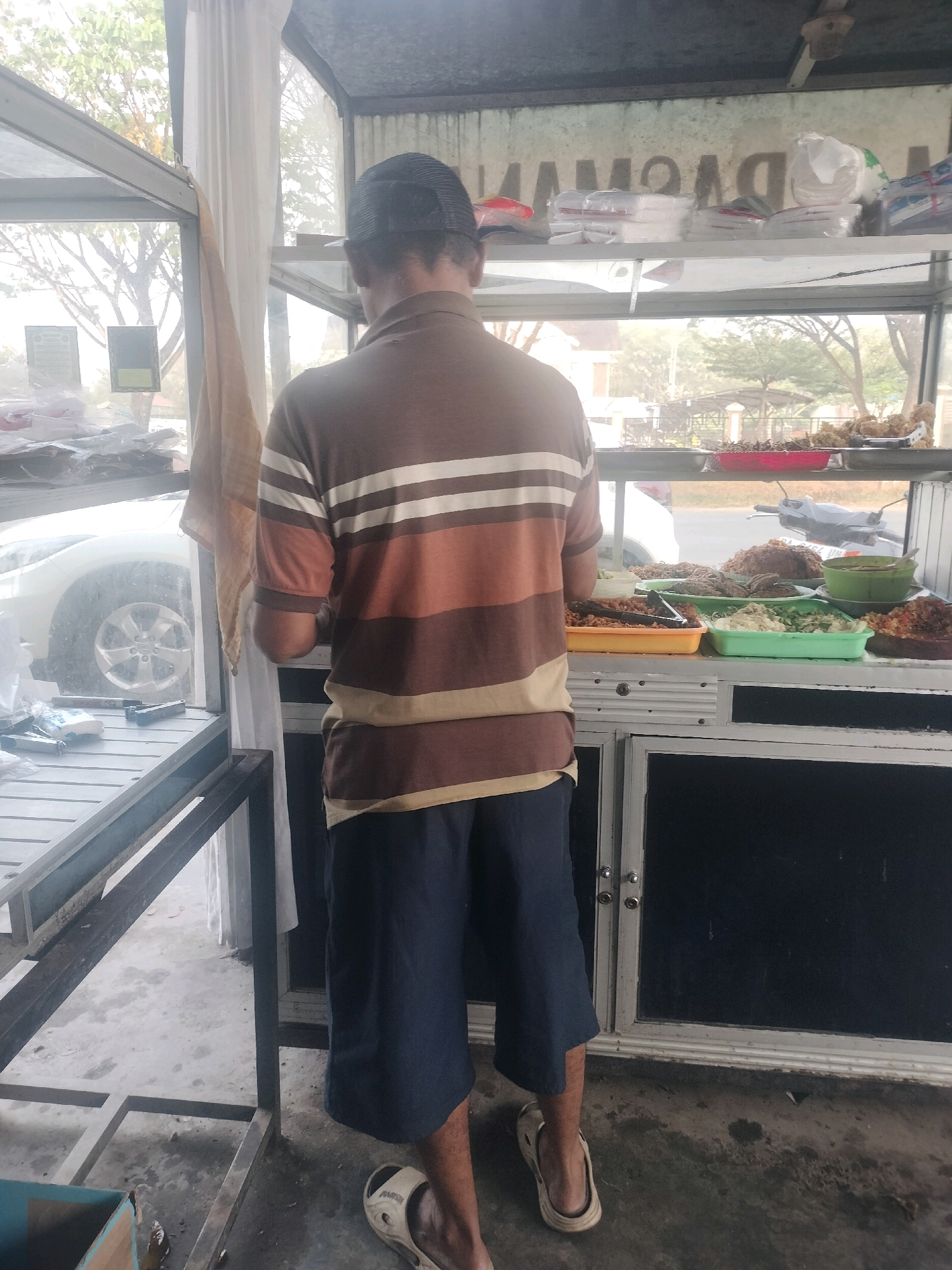 A person wearing a striped t-shirt and shorts stands in front of a food stall in Maburai, Indonesia. The stall has a glass display case filled with various food items, including fried chicken and vegetables. The person is looking at the food, possibly a customer. The background shows a street with a car and a motorcycle, suggesting an outdoor setting. The overall scene depicts a typical street food vendor in Indonesia.