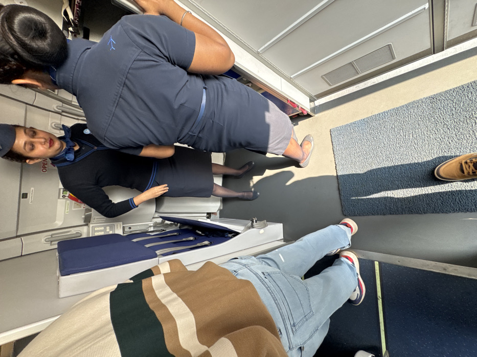 Inside an airplane, two flight attendants in navy uniforms stand near the doorway. One wears a hat and a neck scarf. Facing away, a passenger wears jeans, a striped shirt, and red and white sneakers. A second passenger's brown shoe is visible on a blue carpet. Silver trays, cutlery, and blue headrests are nearby.