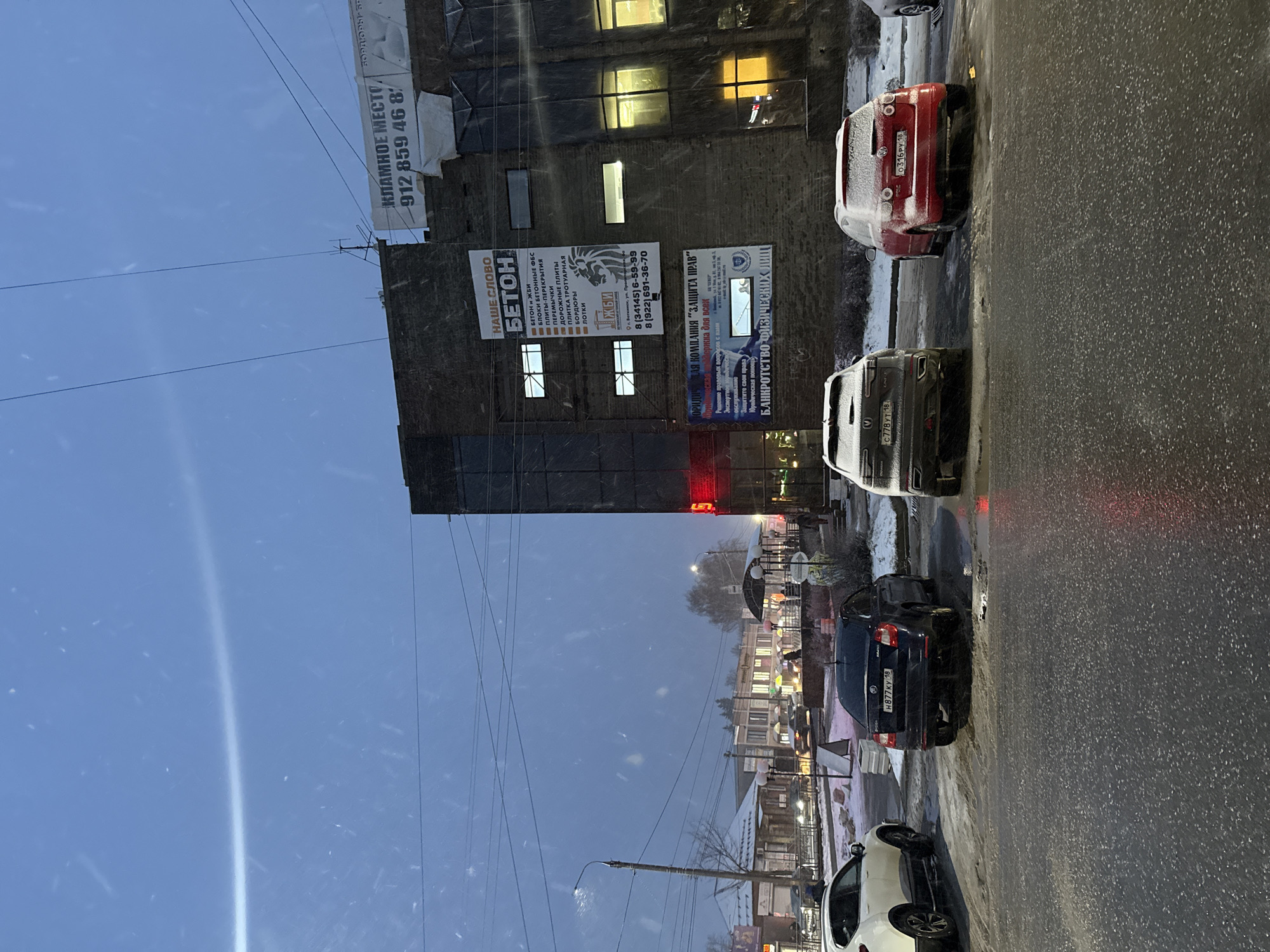 The image depicts a street scene in Votkinsk, Russia, on a snowy day. Several cars are parked along the right side of the street, with a red Volkswagen visible. A tall building lines the left side of the street, covered with advertisements. The advertisements include advertisements for concrete products, a law company ("Zashita Prav"), and a phone number, 912-859-46-8%. There is also a streetlight and power lines above the street. Other buildings are visible in the background, along with some pedestrians. It is dark, and the street is wet.