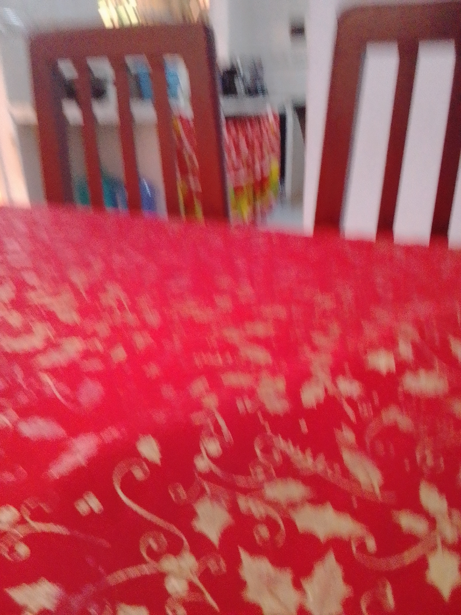 The image shows a red tablecloth with a faint floral pattern draped over a table. In the background, out of focus, are wooden chairs. To the left, the backs of two wooden chairs are visible, with what appear to be colorful objects or bottles behind them. To the right, the backs of two more wooden chairs are visible, with white inserts. The scene is indoors, likely a dining area. There are no people visible in the image. The location is not identifiable from the image itself, therefore, it cannot be determined if the scene is in La Tagua, Venezuela.