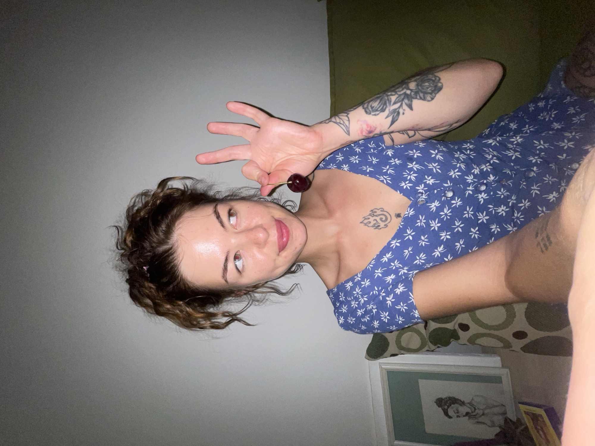 A young woman with curly brown hair pulled up in a messy bun is shown from the chest up, sitting on a couch. She is wearing a blue dress with a white floral pattern. She holds a cherry between her thumb and index finger of her right hand, her other fingers extended. She has tattoos on her chest and left arm. The background is a plain wall and a couch. A framed picture is visible on the left side of the frame. The scene appears to be indoors, in a residential setting. There is no information to indicate the location is in Boreti, Montenegro.