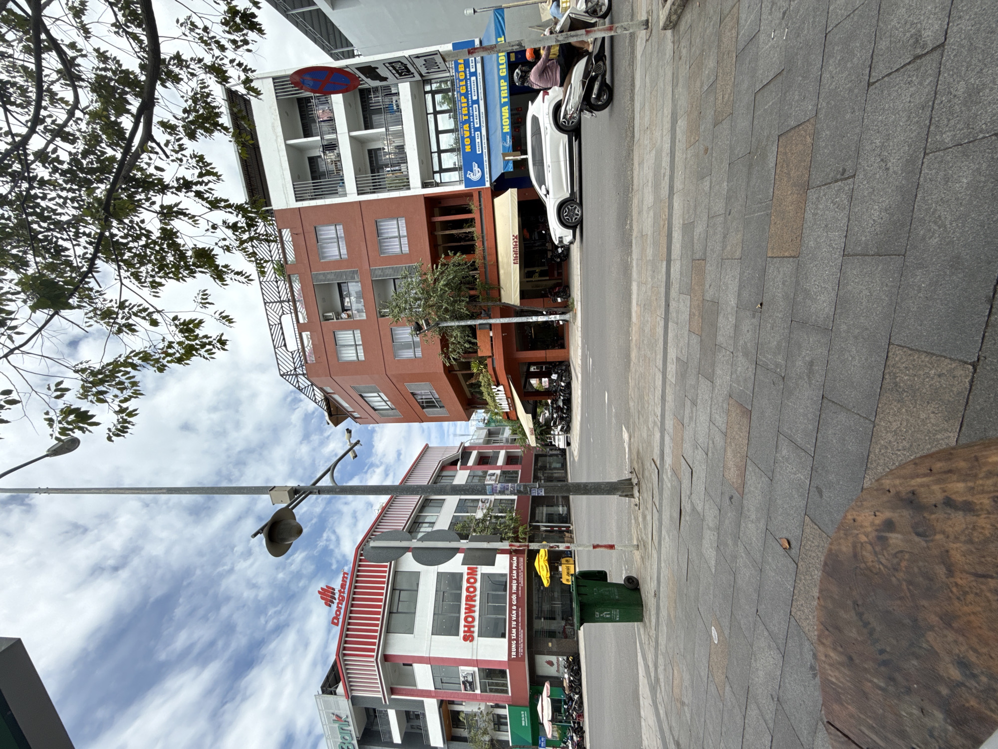 A street scene in Nha Trang, Vietnam, shows a sidewalk in the foreground with a circular wooden table. A paved street stretches into the distance, lined with multi-story buildings. On the left, a white building with red accents features a "Dongtam SHOWROOM" sign. A green trash can stands on the sidewalk. To the right, a reddish-orange building houses "Mamay" and "NOVA TRIP GLOBAL." Several motorcycles are parked along the street and sidewalk. A white SUV and two motorcycles are in motion on the street, one carrying two people. The sky is partly cloudy.