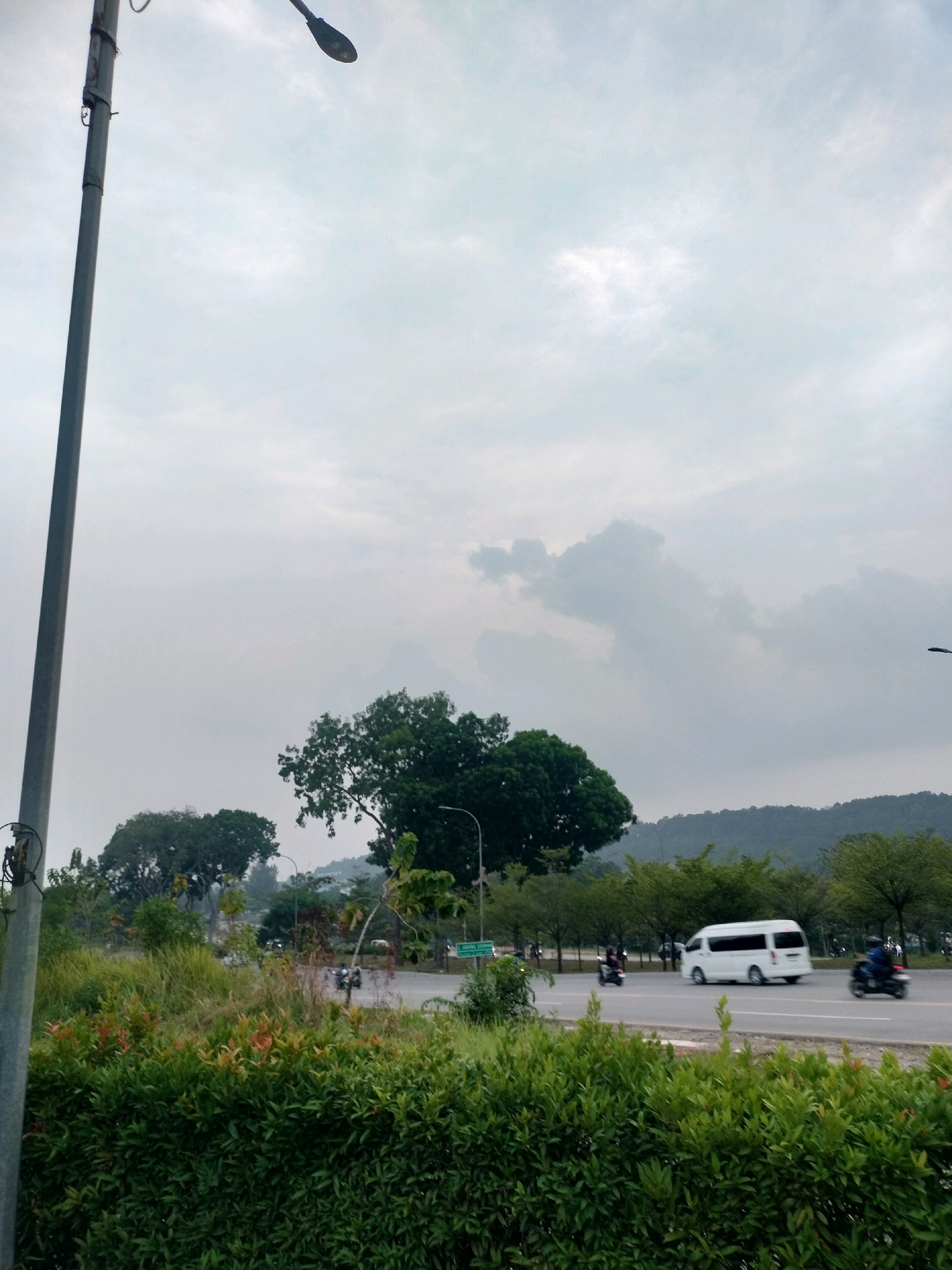 This image depicts a view of a road in Batam, Indonesia, likely on a cloudy day. In the foreground, there are dense green bushes and tall grass. A light pole stands prominently on the left side of the frame. The road, visible in the middle ground, has traffic including a white van and several motorcycles. On the right side of the road, trees line the path, and further in the background, a hilly, tree-covered landscape is visible, with houses or buildings nestled within the hills. The sky is overcast with diffused light.
