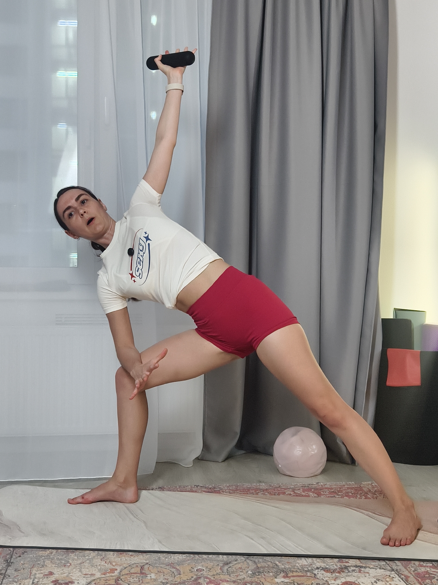 A woman is performing a yoga pose in an indoor setting. She is standing on a yoga mat with her legs spread apart, one arm extended upwards holding a dark cylindrical object, and the other hand resting on her knee. She is wearing a light-colored t-shirt with the word "Sexy" on it, along with red athletic shorts. The background includes a window with white curtains, gray curtains, and yoga blocks stacked against a wall. A pink decorative sphere sits on the mat near her feet. This scene does not contain enough information to determine if this image was taken in the city of Tyumen, Russia.
