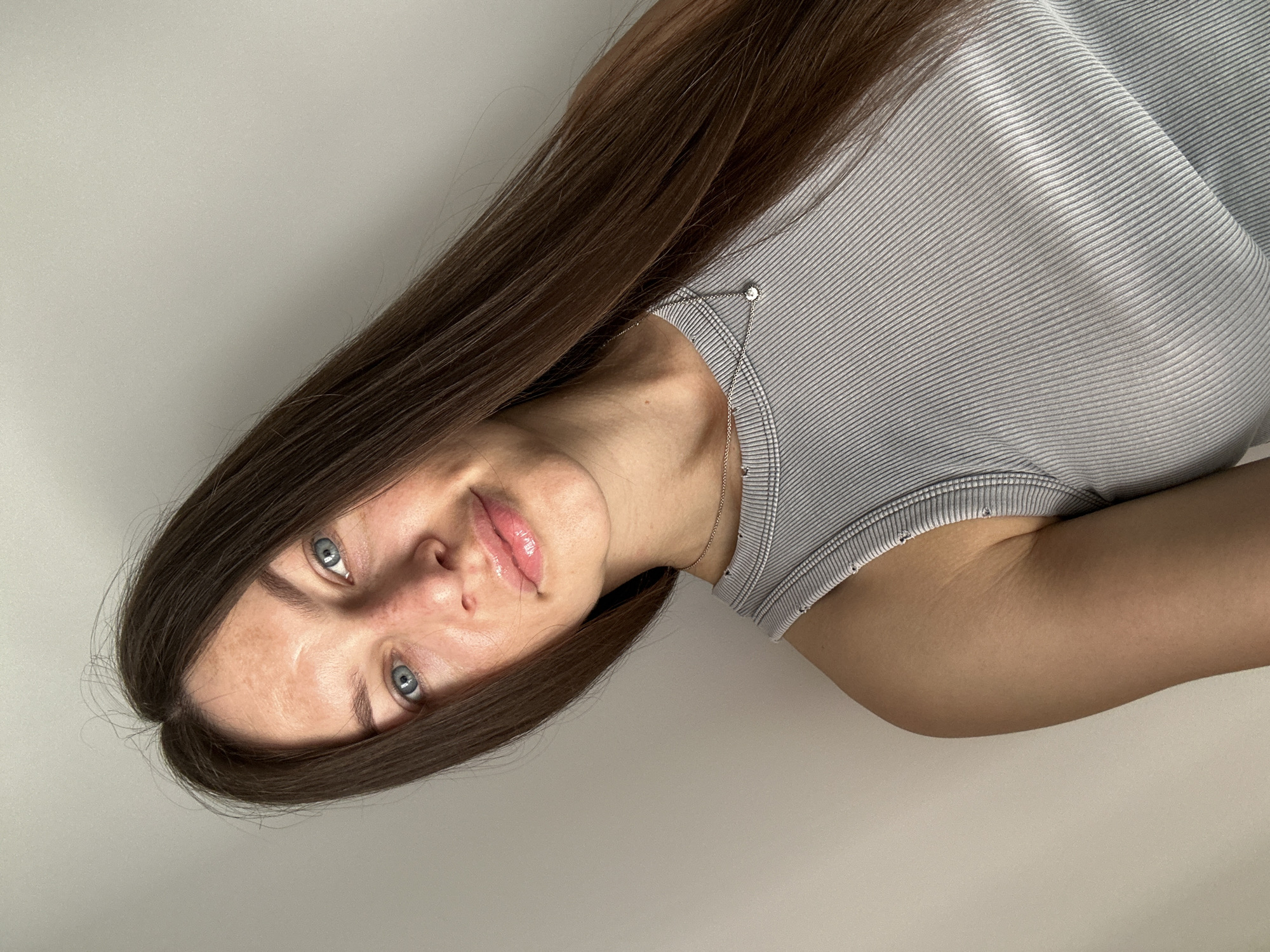 A light-skinned woman with long, dark brown hair is the primary subject of the image. She has striking blue eyes and is wearing a gray, ribbed tank top. A delicate, silver-toned necklace adorns her neck. The background is a plain, off-white surface. The image appears to be taken indoors, and the subject seems to be lying down. There is no indication of the image being in Budva, Montenegro or any city for that matter. The image focuses primarily on the woman's face and upper body.
