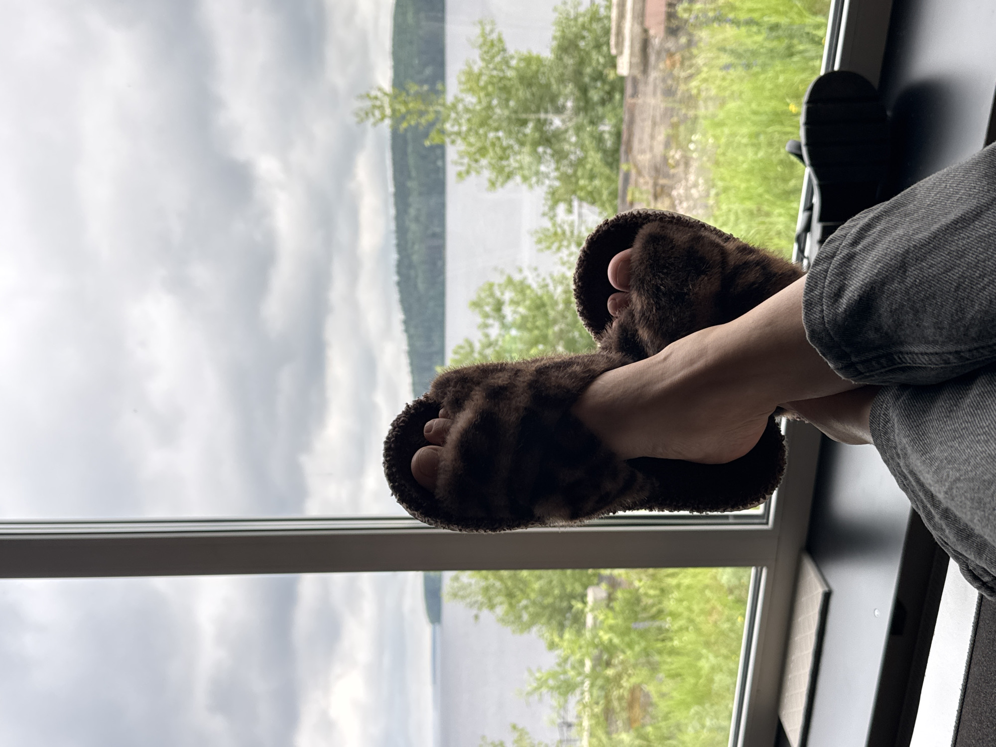 A close-up shot features a person's bare feet, adorned with fuzzy, brown leopard-print slippers, resting in front of a large window. The feet are positioned as if propped up on a surface, with one foot slightly elevated above the other. Beyond the window, a calm body of water stretches out, bordered by a lush, green landscape with trees. The sky above is overcast with gray clouds, suggesting a temperate climate. The scene appears to be indoors, with a dark surface visible beneath the window and a portion of gray fabric, likely clothing, in the foreground. The image evokes a sense of relaxation and tranquility, with the natural scenery offering a peaceful backdrop. The location is identified as Chaikovsky, Russia, a city situated on the Kama River, which aligns with the visual elements of a large body of water and natural surroundings.