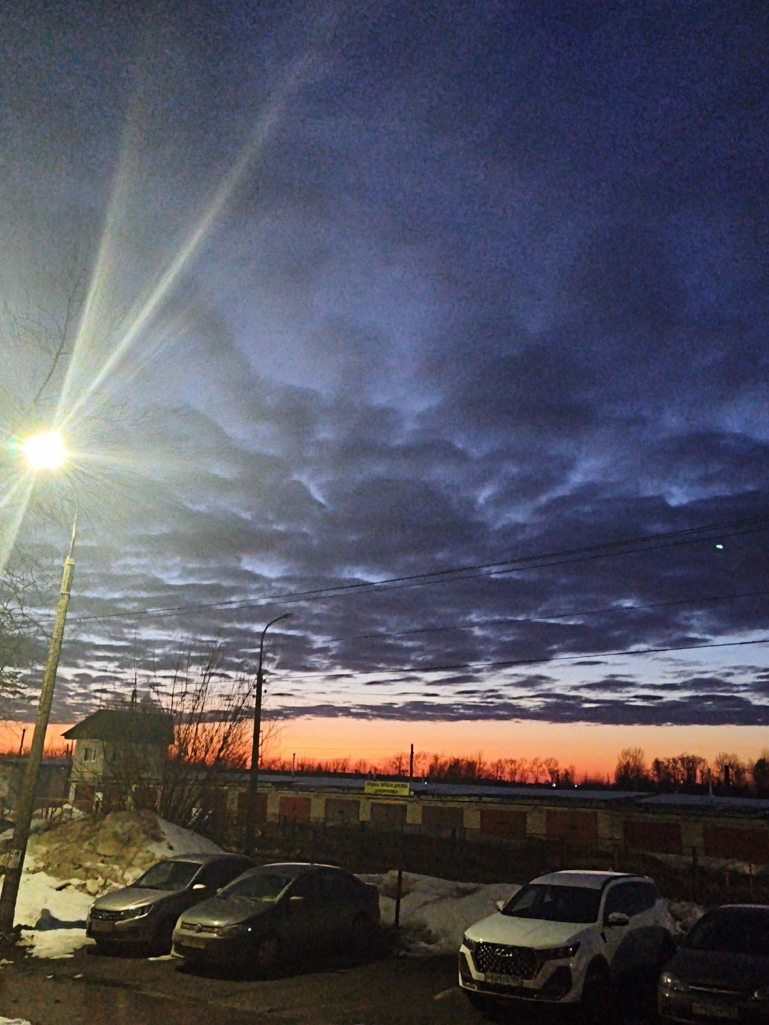 The image depicts an outdoor scene in Chaikovsky, Russia, during dusk or dawn, characterized by a vivid orange and red horizon transitioning to a dark blue-purple sky with scattered clouds. In the foreground, a bright street light mounted on a wooden pole on the far left illuminates bare tree branches and patches of snow on the ground, which also features dark asphalt. Three parked vehicles are visible in the mid-ground: a light-colored sedan on the left, a darker sedan to its right, and a white SUV positioned further right, with the front of a fourth dark vehicle partially visible on the far right. Behind these vehicles, a small, light-colored house or utility building with a dark roof sits on an elevated, snow-covered bank. To the right of this building and extending across the middle distance is a long row of brick or rendered garage-like structures with dark roofs. Overhead, utility lines stretch across the frame, with a dark, unlit street light on a metal pole visible to the right of the frame's center. No individuals or active interactions are present.