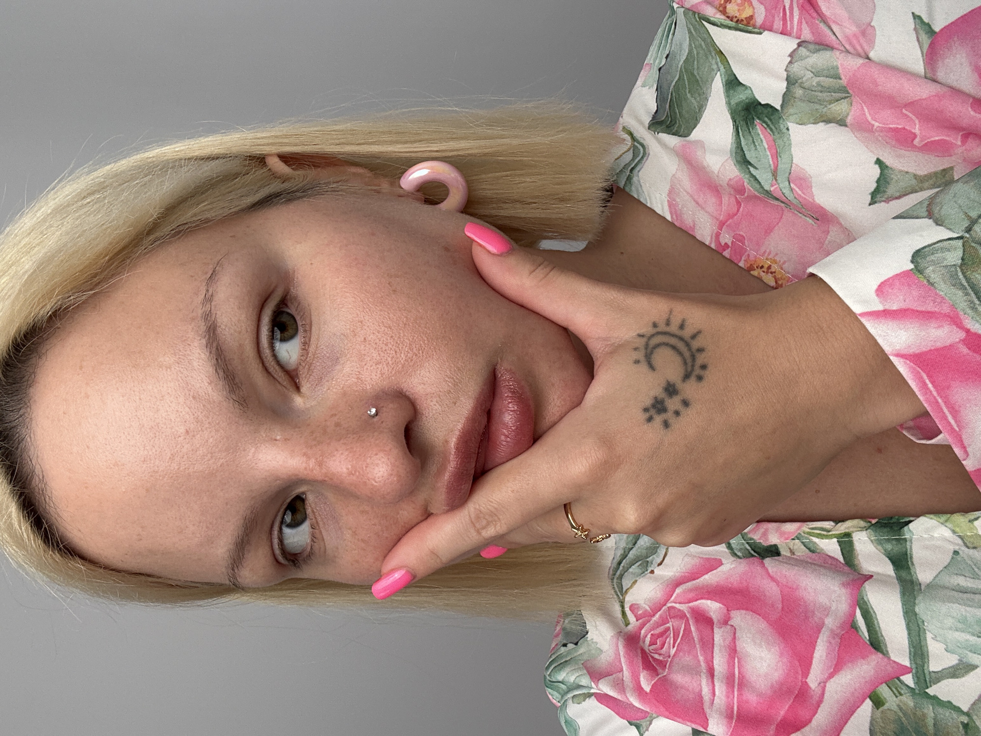 A close-up portrait shows a person with blonde, short hair and fair skin, oriented with their head tilted upwards. They are wearing a white top with a vibrant floral pattern of large pink roses and green leaves. Their left hand, with bright pink nail polish and a gold-colored ring featuring a small star, rests gently against their cheek and jawline. A distinctive black tattoo on the back of this hand depicts a crescent moon with a face, accompanied by a cluster of three small stars.
The person has a small, sparkling stud piercing on their nose and a glossy, C-shaped pink ear gauge in their right ear. Their eyes are dark and appear to be looking towards the camera or slightly above it. The expression is neutral or subtly contemplative. The background is a plain, light grey wall, suggesting an indoor setting.