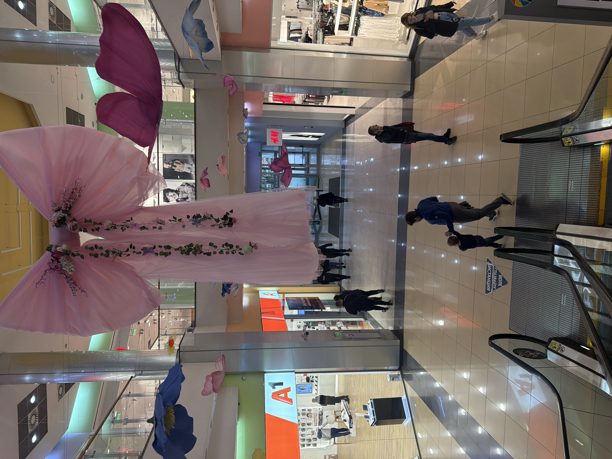 The image shows the interior of a shopping mall in Pleven, Bulgaria. An oversized, decorative pink dress with floral embellishments hangs suspended from the ceiling as the central focal point. Decorative butterflies and flowers are also suspended. Several pedestrians are walking through the mall, near stores like H&M and A1. There are escalators on the right side of the frame. The floor is tiled with a light-colored material and the lighting is bright.