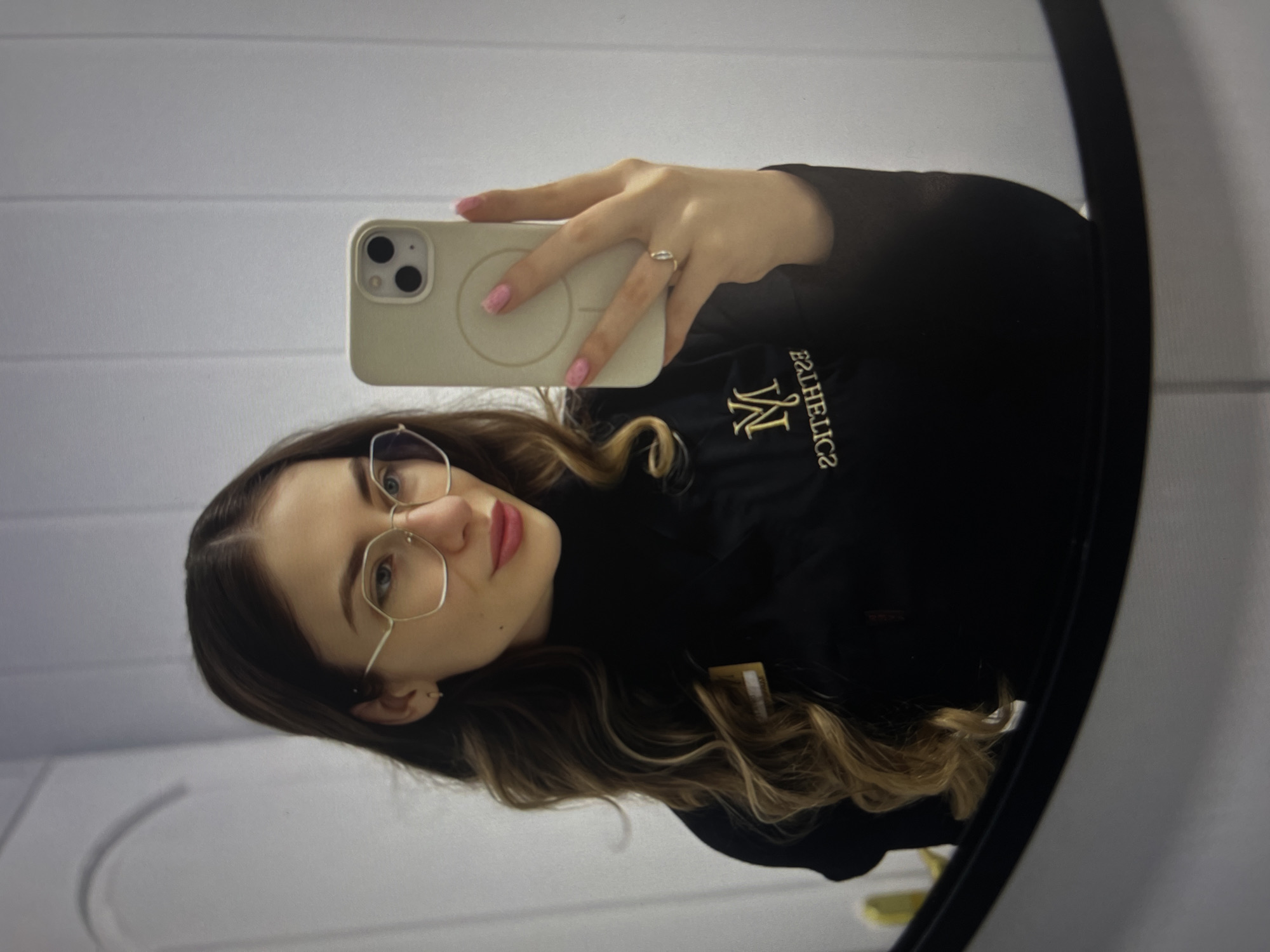 A young woman is taking a selfie in a mirror. She is holding a smartphone in her right hand. Her hair is long, wavy, and dark with light blonde highlights. She is wearing round silver glasses, a black turtleneck sweater with gold writing on the left chest. She has a ring on her right ring finger and her fingernails are painted pink. The background shows a white wall. The photo is taken indoors in Unknown, Unknown.