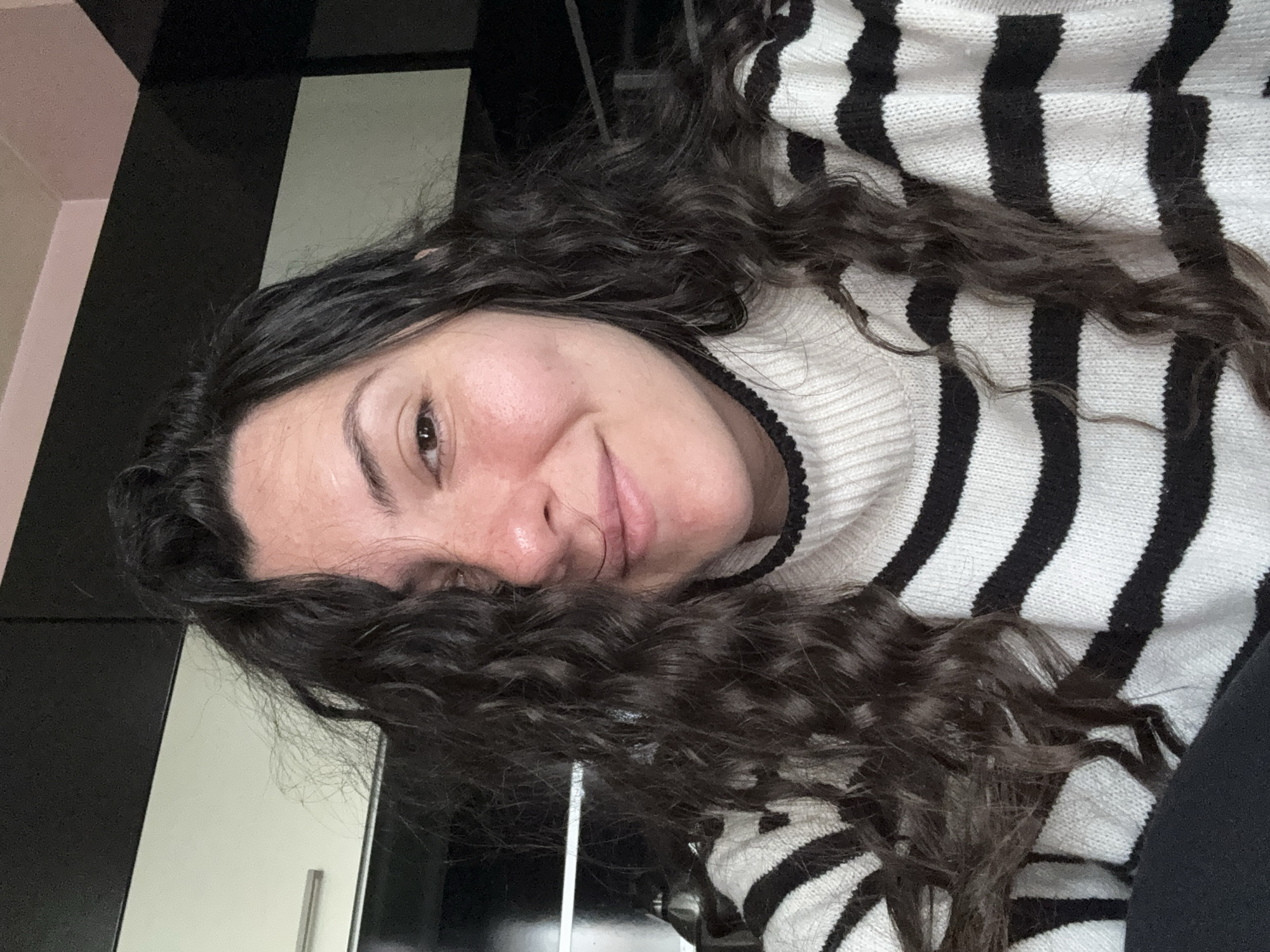A young woman with long, curly brown hair is lying down, possibly on a bed or couch, indoors. She is wearing a black and white striped sweater with a turtleneck. Her face is close to the camera, and she has a slight smile. The background includes a black and white surface, possibly a piece of furniture or a wall, and what appears to be a closed, light-colored door or cabinet. Location is unknown.
