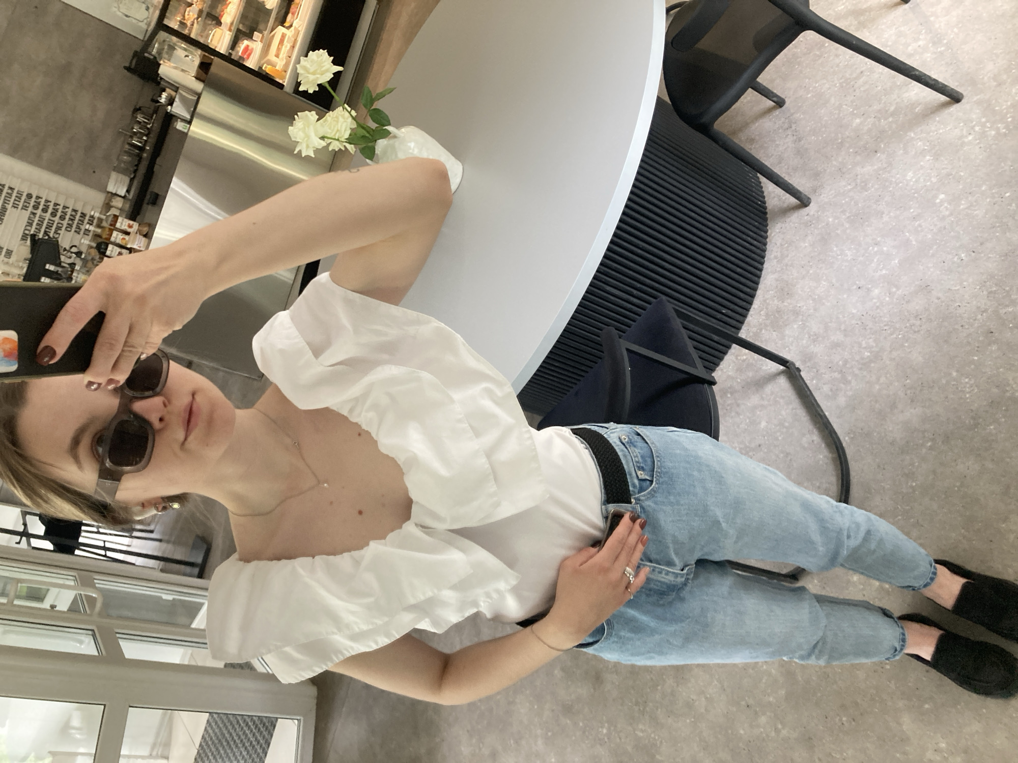 A young adult female is holding a smartphone and taking a selfie in an indoor cafe or restaurant. She is wearing sunglasses, a white off-the-shoulder top, light blue jeans with a black belt, and black shoes. She is standing next to a round white table with a vase of white roses on it. The cafe appears to have modern decor with black chairs and a metal display case with food items in the background. The exact location within the city of Chaikovsky, Russia, cannot be determined from this image.