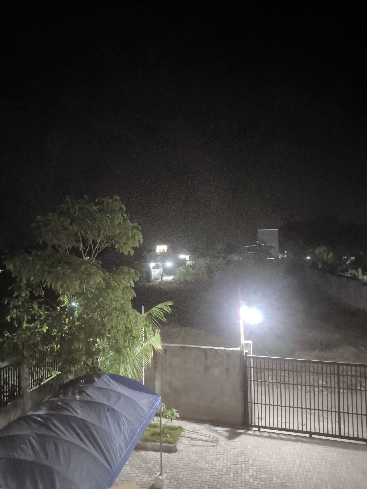 This is a night-time outdoor shot captured from a slightly elevated position, looking out over a landscape. In the foreground, a blue tarp is spread over an unseen object, and to its right, a metal fence encloses a paved area. Beyond the fence, a concrete wall forms a barrier, and a bright light illuminates a flagpole with a red and white flag. The middle ground shows a hillside with some vegetation, and scattered lights indicate buildings or homes. A prominent tree with green foliage stands on the left side of the frame, partially obscuring the view of the distant structures. The background is a dark, starless sky. The scene appears to be in a residential or semi-rural area, possibly a backyard or balcony overlooking a neighborhood.