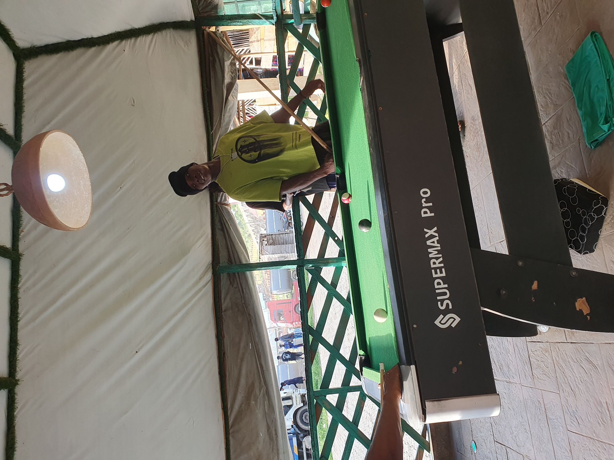 The image depicts a person playing billiards inside a tent-like structure. The person is wearing a light green t-shirt and a black cap, and is holding a billiard cue, aiming at a ball on the green felt table. The table has the branding "SUPERMAX Pro" visible on its side. Several billiard balls are scattered on the table.
Outside the tent, through a green latticed opening, a street scene is visible with several vehicles, including trucks and cars. Some people are also seen in the background, appearing to be walking or standing. The lighting suggests it is daytime. The tent has white fabric walls and a floor covered with artificial turf. A decorative lamp made from a gourd hangs from the ceiling. The overall setting appears to be an informal outdoor area, possibly a recreational space or a market.