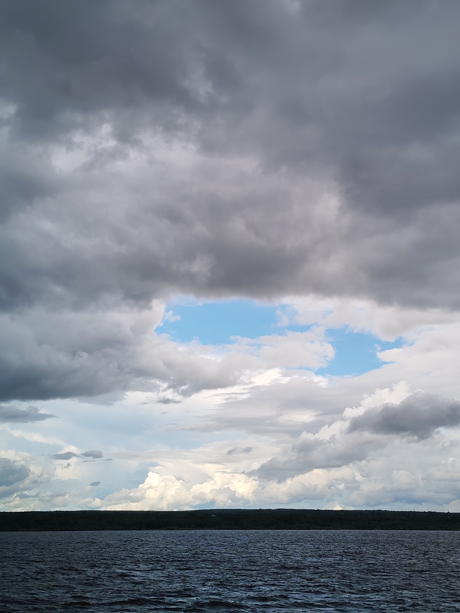 A wide-angle, landscape view captures a dark, choppy body of water in the foreground, reflecting the overcast sky above. A dark, dense tree line runs horizontally across the middle ground, separating the water from the sky. The sky is dominated by heavy, gray clouds, with a small patch of bright blue sky visible near the center. White, puffy clouds are scattered throughout the lower portion of the sky, creating a contrast with the darker clouds. The scene appears to be a natural landscape, possibly a lake or a large river, with no signs of human activity or structures visible. The overall impression is one of a dramatic and moody atmosphere.