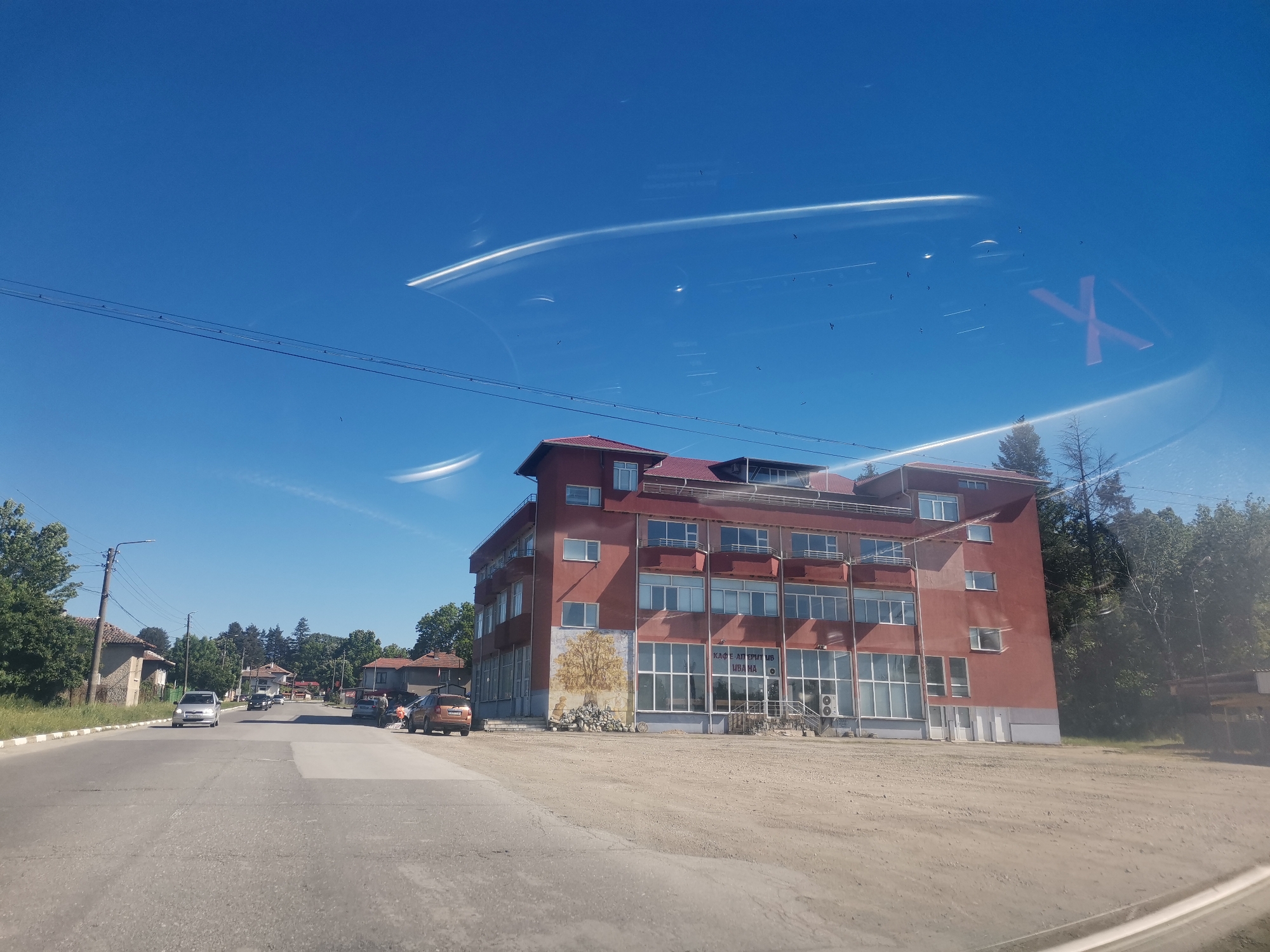 The image shows a street scene in the village of Mirovo, Bulgaria. A three-story reddish-brown building with a red roof and many windows is located on the right side of the road. The building has balconies on the second and third floors. A mural depicting a tree is visible on the side of the building. In front of the building is a dirt area. The road is asphalt and runs horizontally across the image. There are several cars parked or driving on the road. On the left side of the road is a mix of vegetation and buildings. The sky is clear and blue, with reflections on the windshield. Power lines run along the top of the frame. There are birds in the sky.