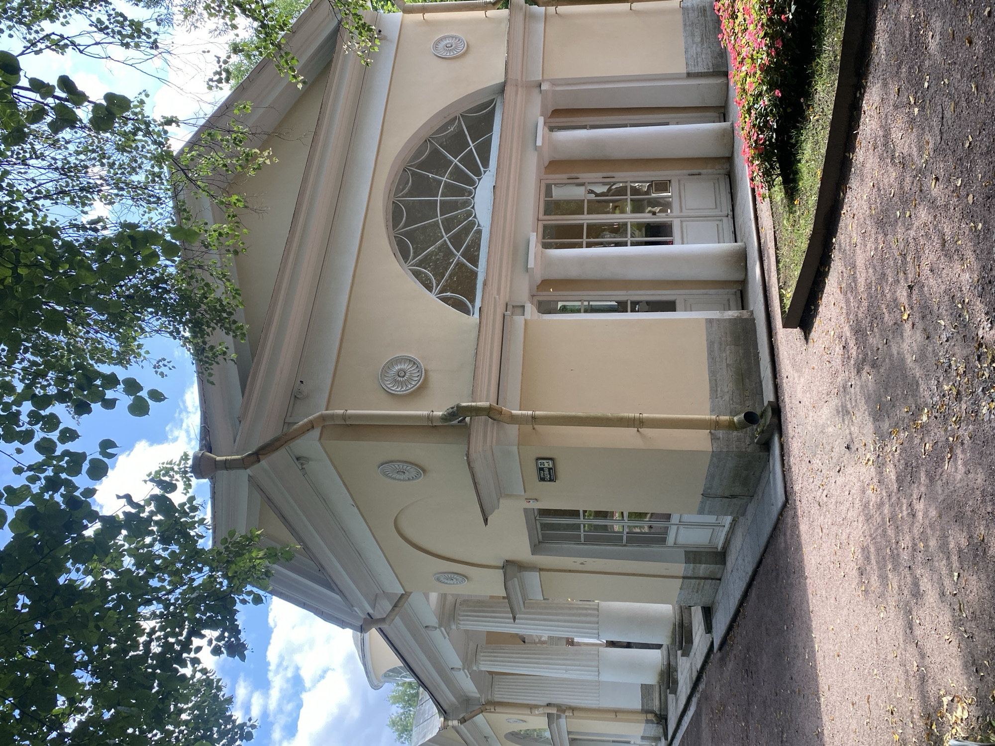The image shows a light yellow classical building with white columns and a large arched window. The building is situated in a park-like setting with gravel paths, trees, and a flower bed. A sign with Russian text reading "Садовая 20" is visible on the wall. The building appears to be part of the Pavlovsk Palace complex in Pavlovsk, Russia. The sky is partly cloudy and the scene is illuminated by sunlight, casting shadows on the ground. No people or animals are visible in the image.
