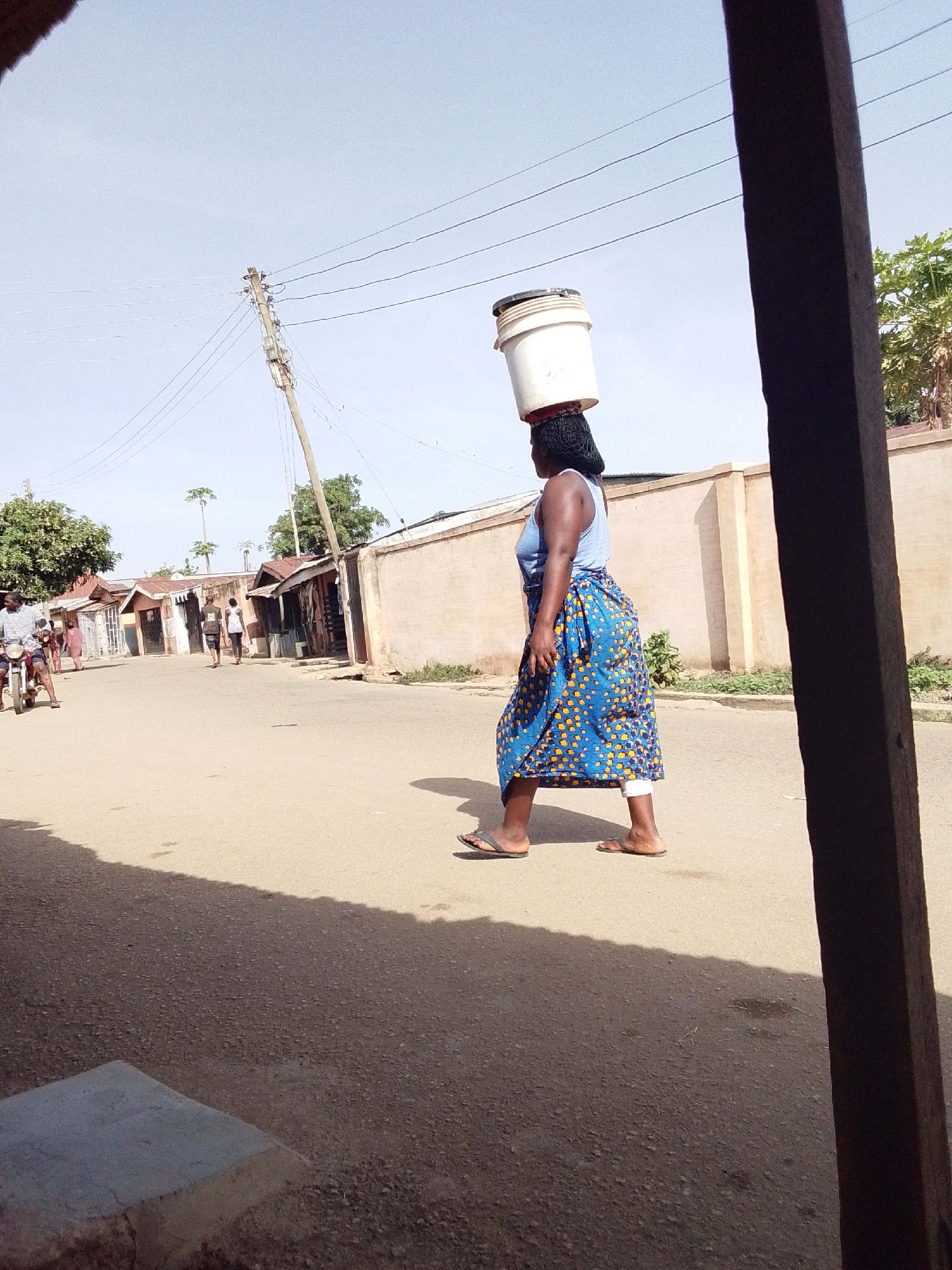 The image captures a daytime scene on a street in Jos, Nigeria. The primary subject is a woman walking from right to left across the frame, balancing two stacked white buckets on her head. She is wearing a light blue tank top, a long blue skirt with yellow patterns, and flip-flops. Her hair is dark and styled, likely forming a base for the buckets. She has a bandage or wrap on her lower right leg.
In the background, the street appears to be a mix of asphalt and dirt, stretching into the distance. On the left side of the road, a man is seated on a motorcycle. Further down the street, two other individuals are walking away from the viewer. The roadside is lined with modest, single-story buildings and walls, some appearing to be houses or small businesses, with corrugated metal roofs visible on several structures. A tall power pole with numerous power lines cuts across the upper left part of the sky. The sky is clear and bright, suggesting sunny weather. A dark wooden beam is visible in the extreme foreground on the right, partially obscuring the view. There is no visible text in the image.