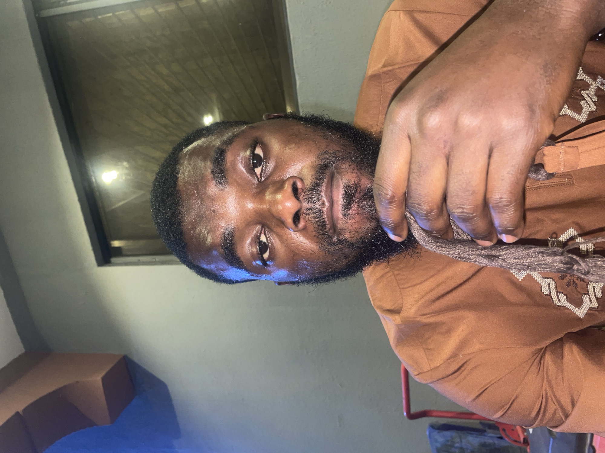 A man with dark skin and a full beard is seen in the image, wearing a brown traditional garment with embroidered details. He appears to be indoors, with a window visible in the background. The lighting suggests it may be evening or a dimly lit space. The context of Jos, Nigeria, is not evident from the image itself.
