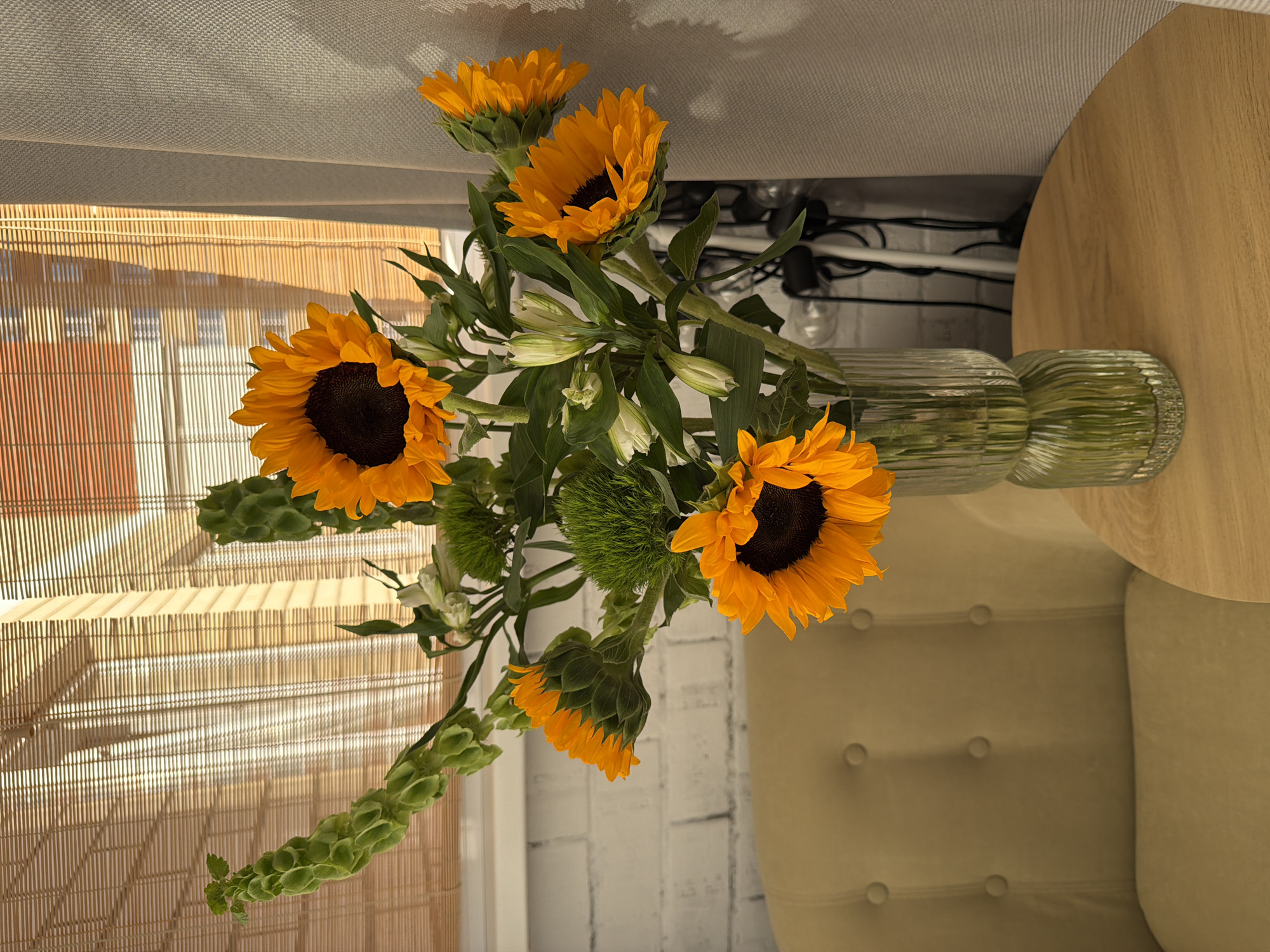 A bouquet of sunflowers, lilies, and green bells of Ireland rests in a clear glass vase. The vase is positioned on a wooden circular table adjacent to a tufted light yellow chair. A bamboo window shade is visible in the background on the left side of the image. Behind the flowers, a white brick wall is partially visible. An electrical cord is lying on the floor. The image is oriented sideways. The location is Unknown, Unknown.