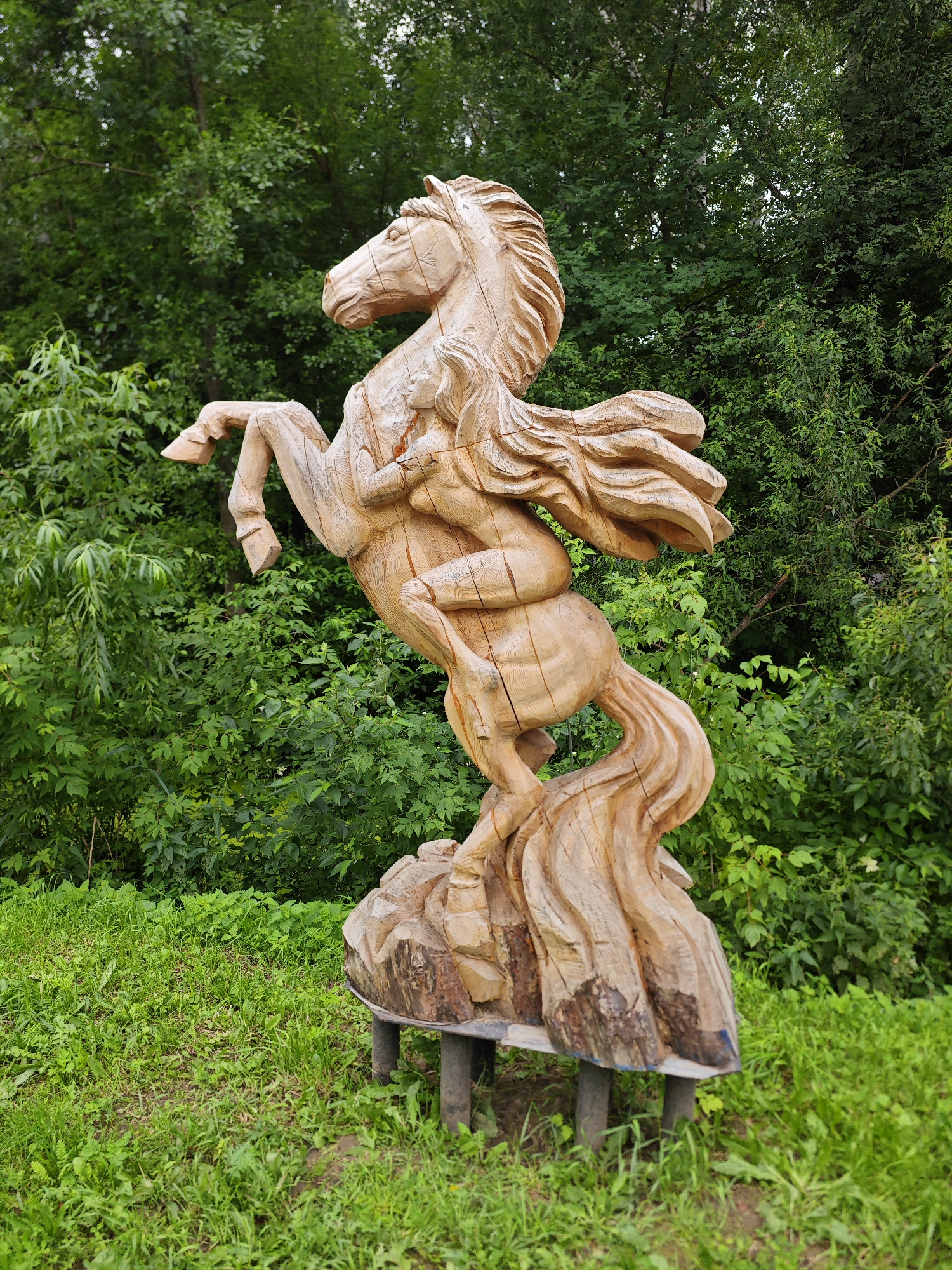 A wooden sculpture of a winged horse is displayed outdoors on a grassy area with a backdrop of dense green foliage. The sculpture depicts the horse rearing up on its hind legs, with its front legs lifted and its wings spread. The wood appears to be roughly hewn, with visible chisel marks and natural wood grain. The sculpture is mounted on a wooden base supported by dark metal legs, which are partially visible on the grass. The environment suggests a park or natural setting. The sculpture is located in the city of Yoshkar-Ola, Russia, in the Mari El Republic.