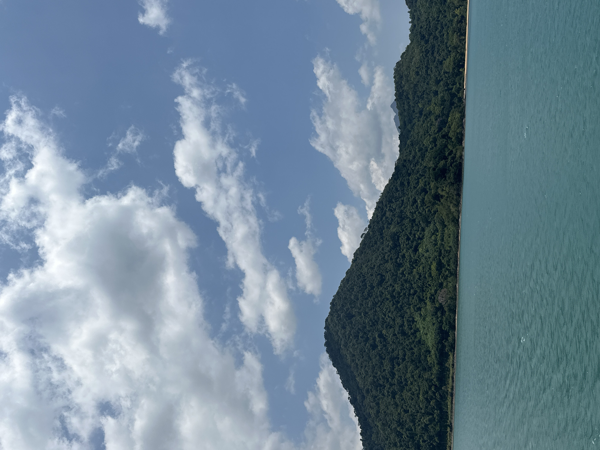 The image shows a landscape in Baan Klong Son, Thailand. On the right, there's a turquoise body of water, possibly the sea. Adjacent to the water is a mountain densely covered with green trees. The sky above is blue with scattered white clouds.