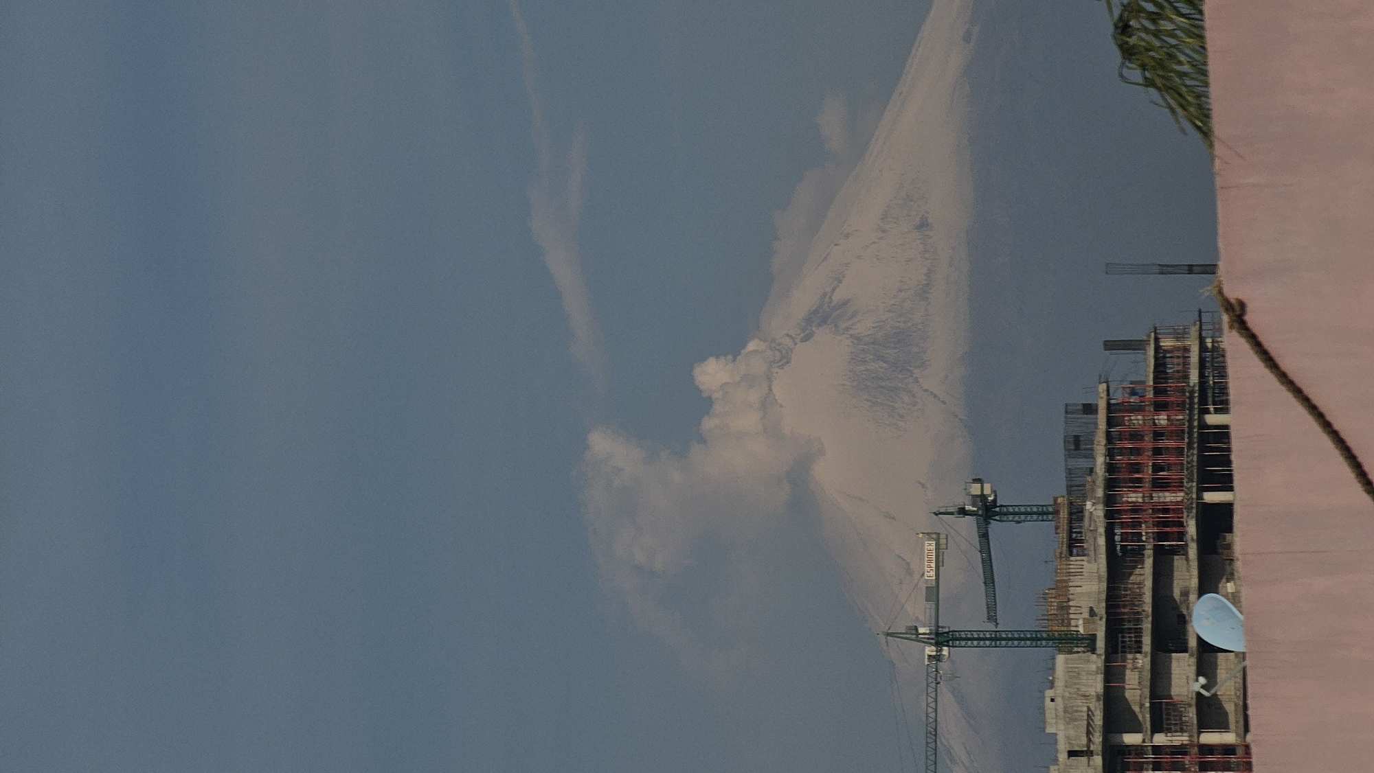 The image depicts a construction site in Puebla, Mexico, situated against a backdrop of the city and a volcano. The scene features high-rise buildings under construction, characterized by scaffolding and cranes. The sky is partly cloudy.