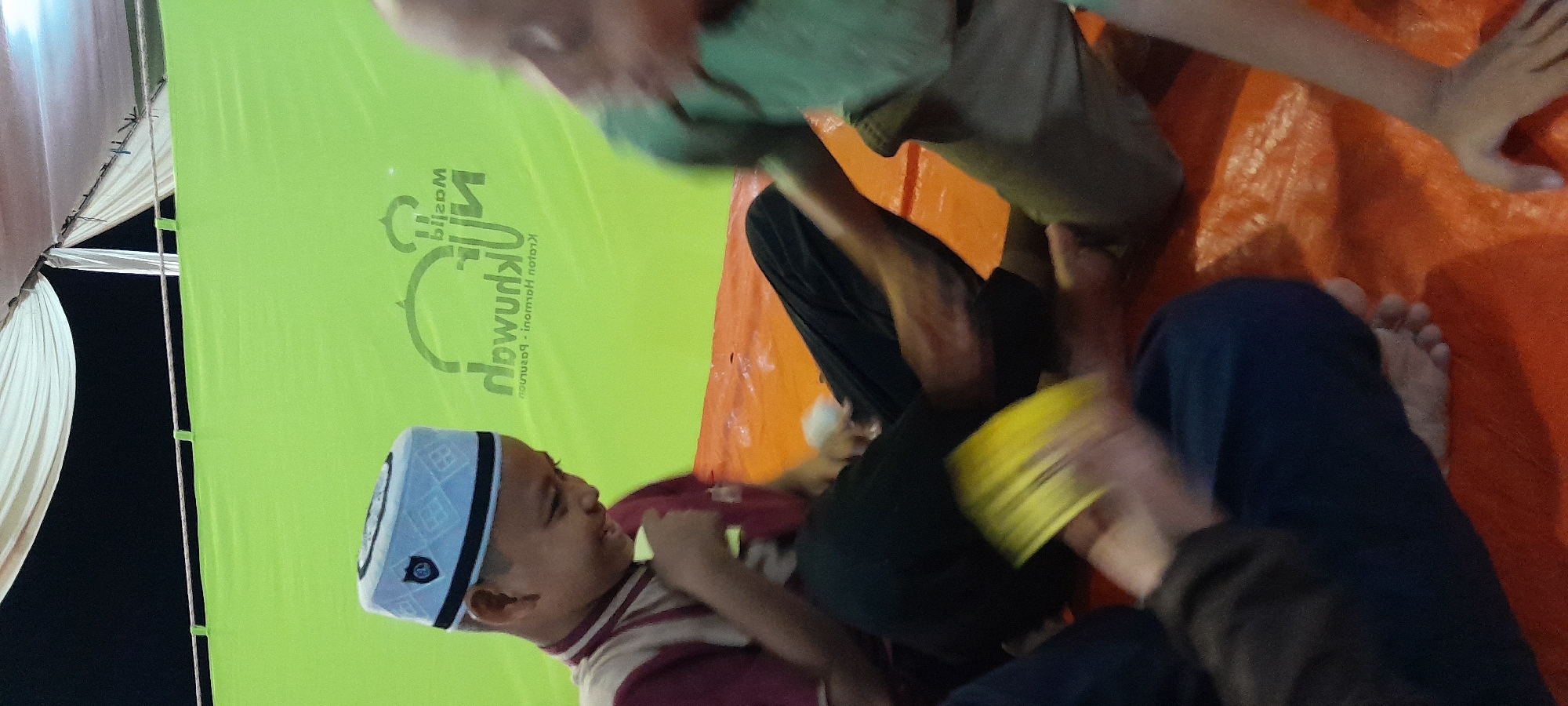 The image captures an indoor scene, likely a community gathering or event, set against a bright green backdrop displaying a logo with Arabic text. In the foreground, a young boy wearing a white kufi cap and a maroon and beige striped shirt is seated on an orange surface, smiling and holding a yellow object. He is looking down and to his right. To his right, another child, partially visible and blurred, is also seated and appears to be interacting. Other individuals' legs and feet are visible in the lower portion of the frame, suggesting multiple people are present and seated on the orange surface. The overall lighting suggests an evening or indoor setting. The logo on the green backdrop appears to be for "Masjid Al-Muhibbin - Krorton Pasuruan - Inormoh Nofor". This suggests the location is in or related to the Pasuruan region of Indonesia, possibly Bendungan, which is a subdistrict in East Java, Indonesia, known for its community and religious activities.