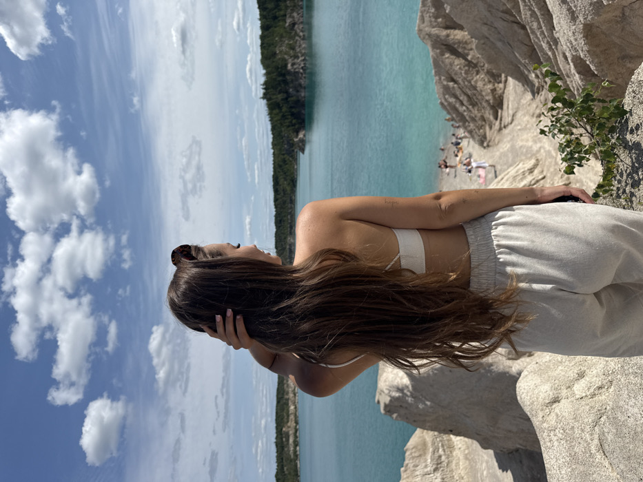 The image presents an outdoor scene in Druzhba, Ukraine, showcasing a body of turquoise water bordered by light-colored, eroded chalk formations and distant forested shores under a clear blue sky with scattered white clouds. A female subject, seen from behind and to her left, stands in the foreground atop an elevated chalk formation, gazing out over the water. She possesses long, dark brown hair, with her right hand positioned near her head. She is attired in a light-colored bikini top and light-toned, loose-fitting trousers, with tortoiseshell sunglasses resting on her head. In the mid-ground, approximately 10-15 individuals are visible on a narrow, light-colored beach directly adjacent to the water. Sparse green vegetation is present on the immediate rocky foreground.