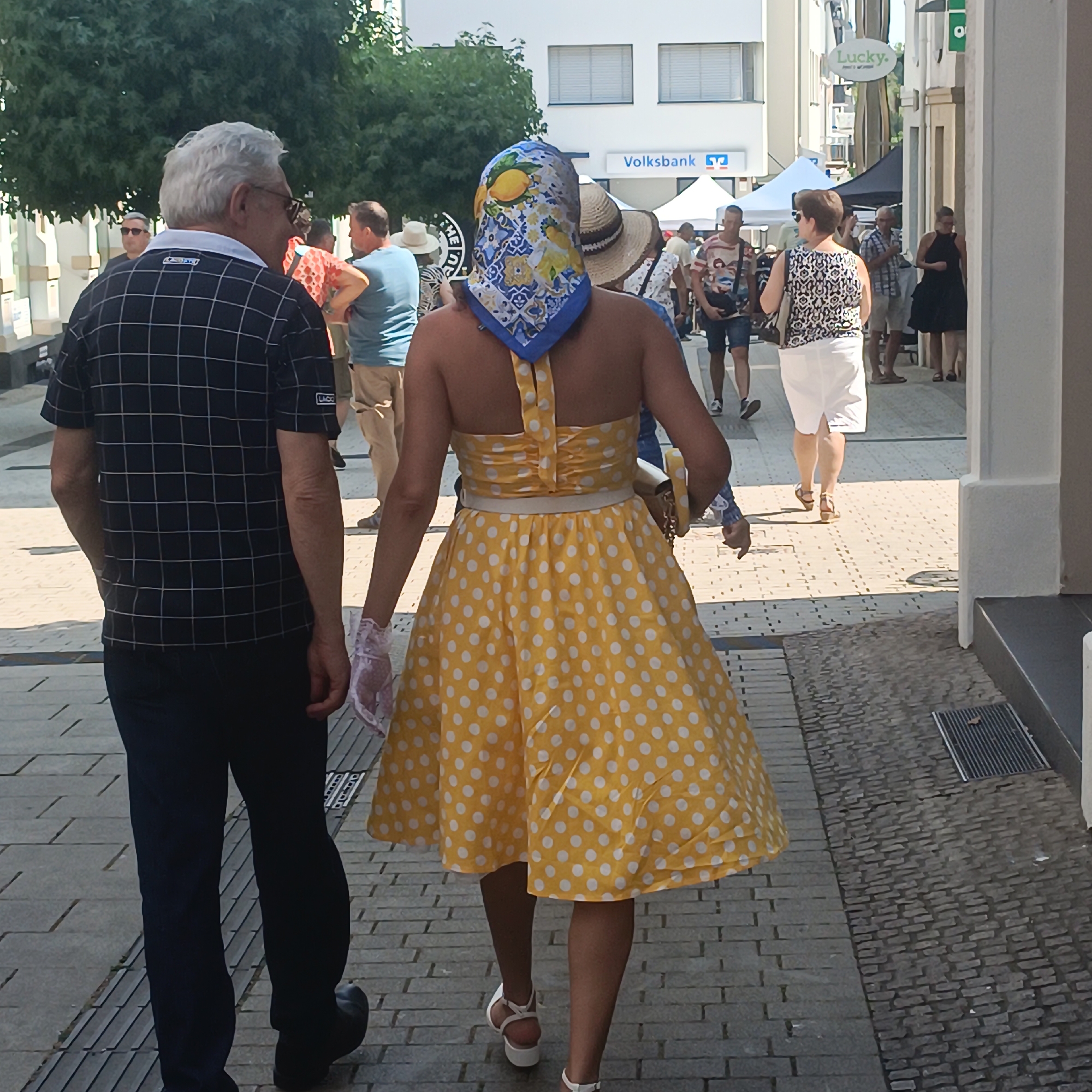 The image depicts a street scene in Bad Nauheim, Germany, likely on a sunny day. In the foreground, a man and a woman are walking away from the camera. The man is wearing a black and white checkered short-sleeved shirt and dark trousers. The woman is wearing a yellow and white polka dot dress and white open-toed sandals. She has a colorful headscarf with a lemon pattern tied around her head. In the background, other people are seen walking, and there are buildings lining the street, some with awnings. A sign for "Volksbank" is visible on one of the buildings. The street is paved with cobblestones.