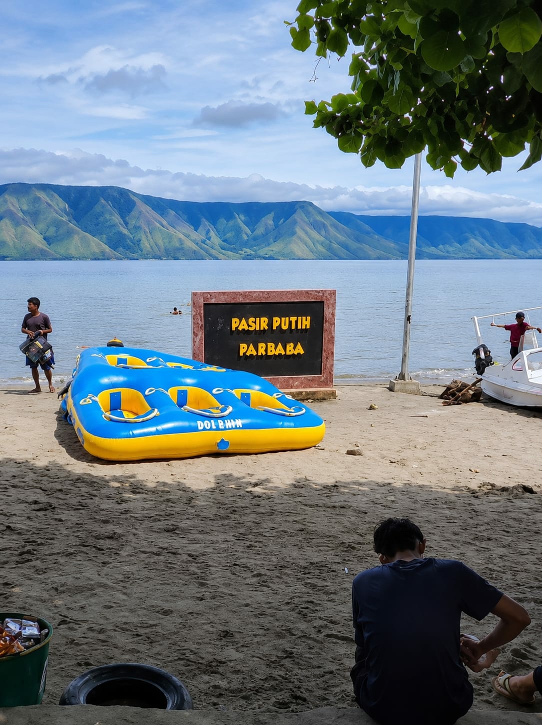 In Situngkir, Indonesia, this image captures a scenic beach with a calm lake and lush green mountains in the background. A blue and yellow inflatable watercraft, labeled "DOLPHIN," rests on the sandy shore. Nearby, a black sign with yellow lettering reads "PASIR PUTIH PARBA." A man in a dark t-shirt stands to the left of the watercraft, holding what appears to be a bag. In the distance, two individuals are visible in the water. To the right, a white boat is docked, and a person in a red shirt stands near it. The foreground shows a man in a dark blue shirt sitting on the sand, facing away from the camera. The scene is set on a bright, cloudy day.