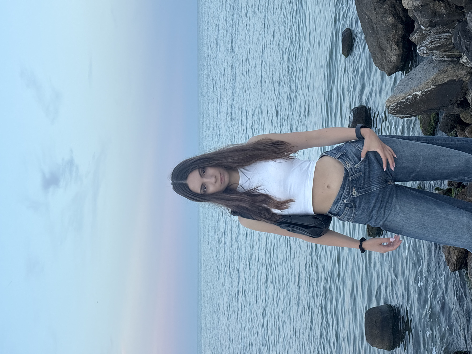 A young woman stands on a rocky shoreline with the sea behind her. The sky is a pale blue with wisps of pink and purple. The woman is wearing a white crop top, blue jeans, and a black purse slung over her shoulder. She has long, dark hair and is looking directly at the camera with a serious expression. Her left hand is in her jeans pocket. The water is calm, with small waves lapping at the shore. There are several large rocks in the water and on the shore. The location is Varna, Bulgaria.