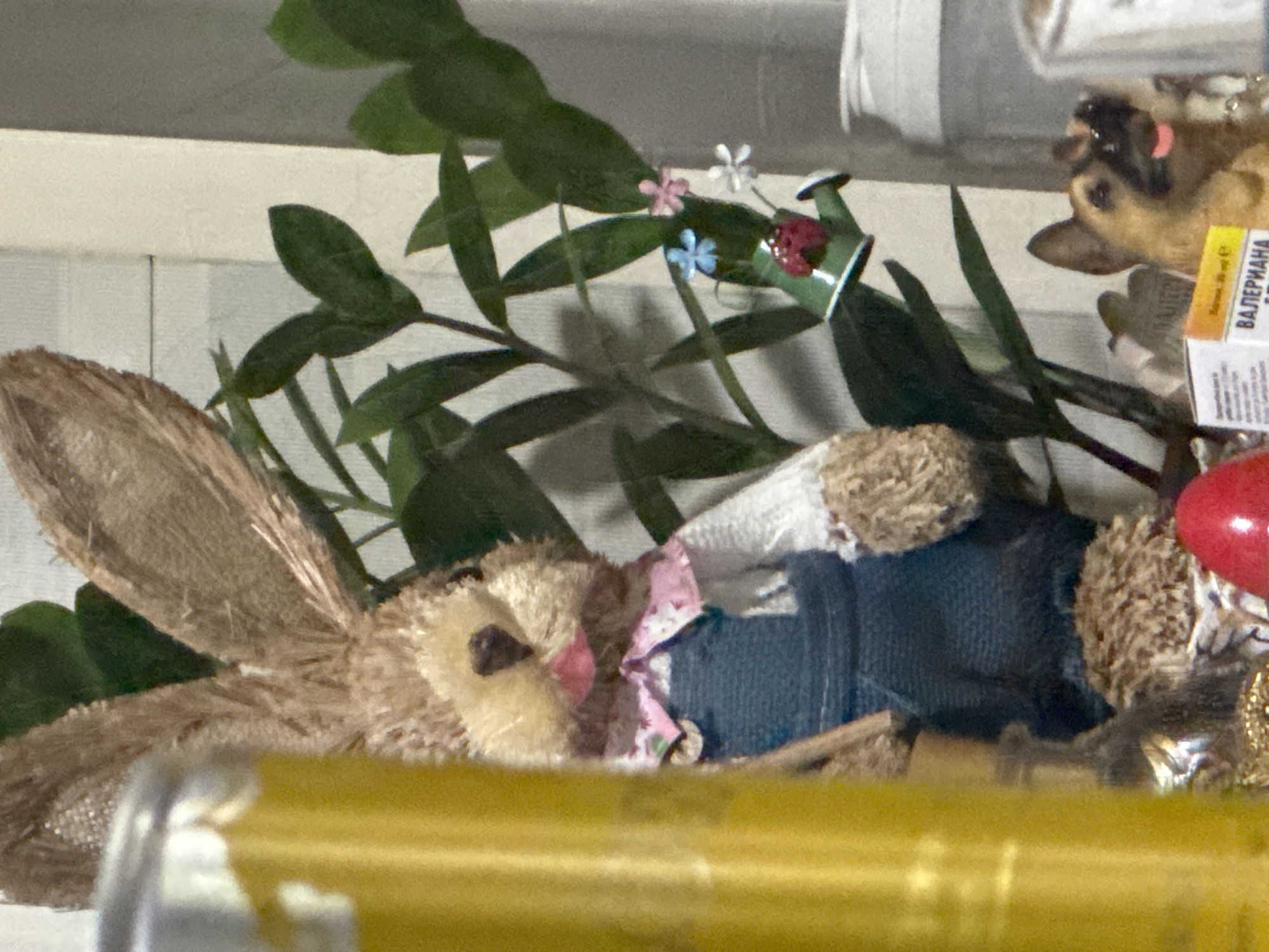 In a close-up indoor shot, a plush Easter bunny toy wearing blue overalls and a pink floral collar stands prominently, partially obscured by a blurred yellow cylindrical object on the left. Behind the bunny, a green leafy plant is visible, adorned with a miniature green watering can featuring a ladybug and tiny flowers. To the right of the plant, a small German Shepherd dog figurine with its tongue out is positioned in front of a box labeled "ВАЛЕРИАНА". The overall scene suggests a decorative display, possibly in a home or shop, with no discernible outdoor elements or specific urban context of Sofia, Bulgaria.