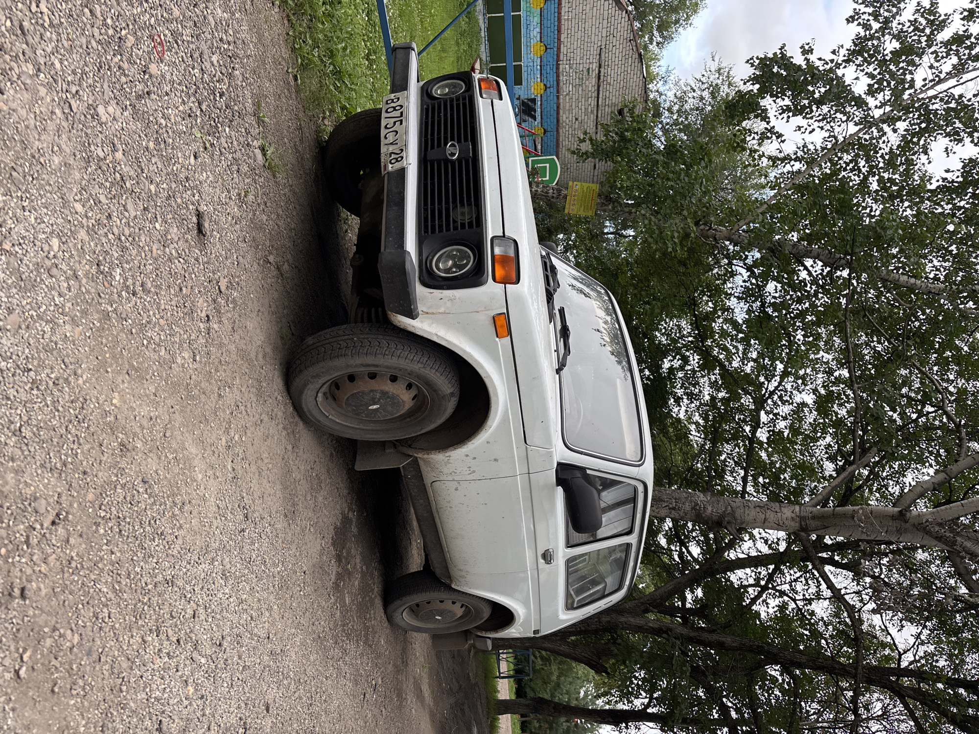 A white, two-door SUV is parked on a gravel path next to a brick building and trees. The vehicle has a license plate that reads "B875CV 28 RUS". The building appears to be a school or community center, with colorful murals on its exterior. Trees with green leaves line the background. The sky is overcast.
