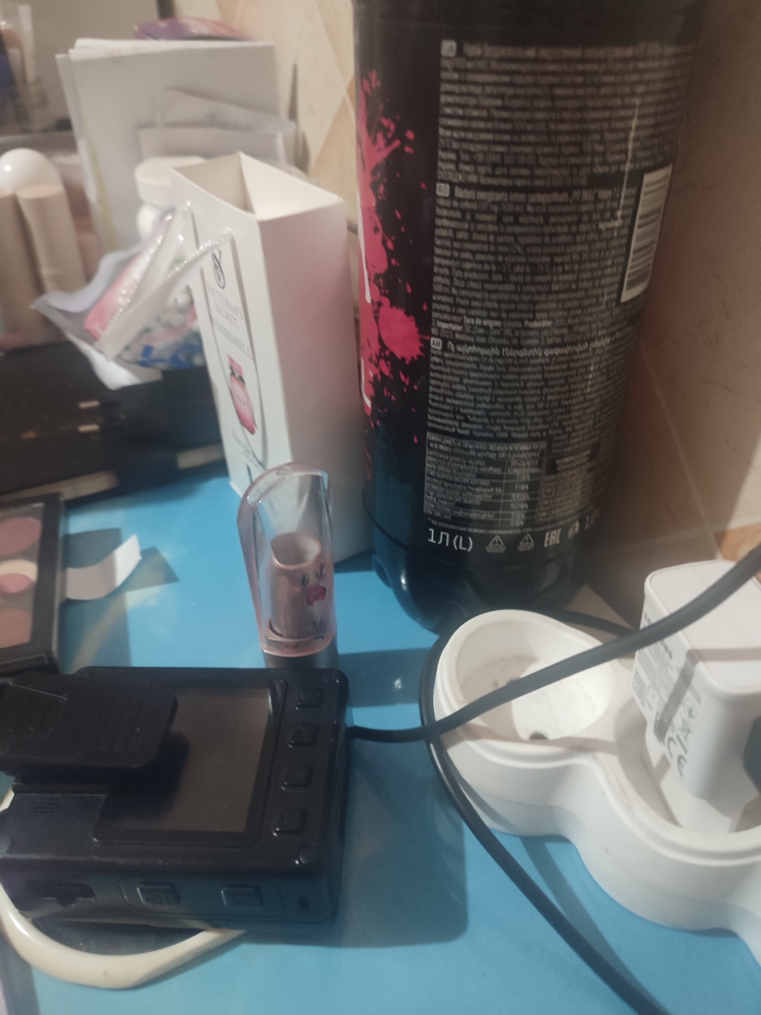 A close-up, eye-level shot displays a collection of items on a light blue surface. In the foreground, a black digital camera is positioned to the left. To the right of the camera, a clear plastic tube, likely a lipstick holder, contains a pink tube with a flower design. Behind these items, a large black bottle with pink splatter patterns dominates the background, with visible text in various languages on its label. Further back, a white gift bag with a pink accent and a visible logo suggests a retail or cosmetic context. Other blurred items, including what appear to be makeup palettes and cylindrical objects, are also present. The lighting is dim, casting shadows and highlighting the textures of the objects. No individuals are visible in the frame. The scene is likely set indoors, on a table or desk, in Chernivtsi, Ukraine, given the presence of text in Armenian on the bottle.