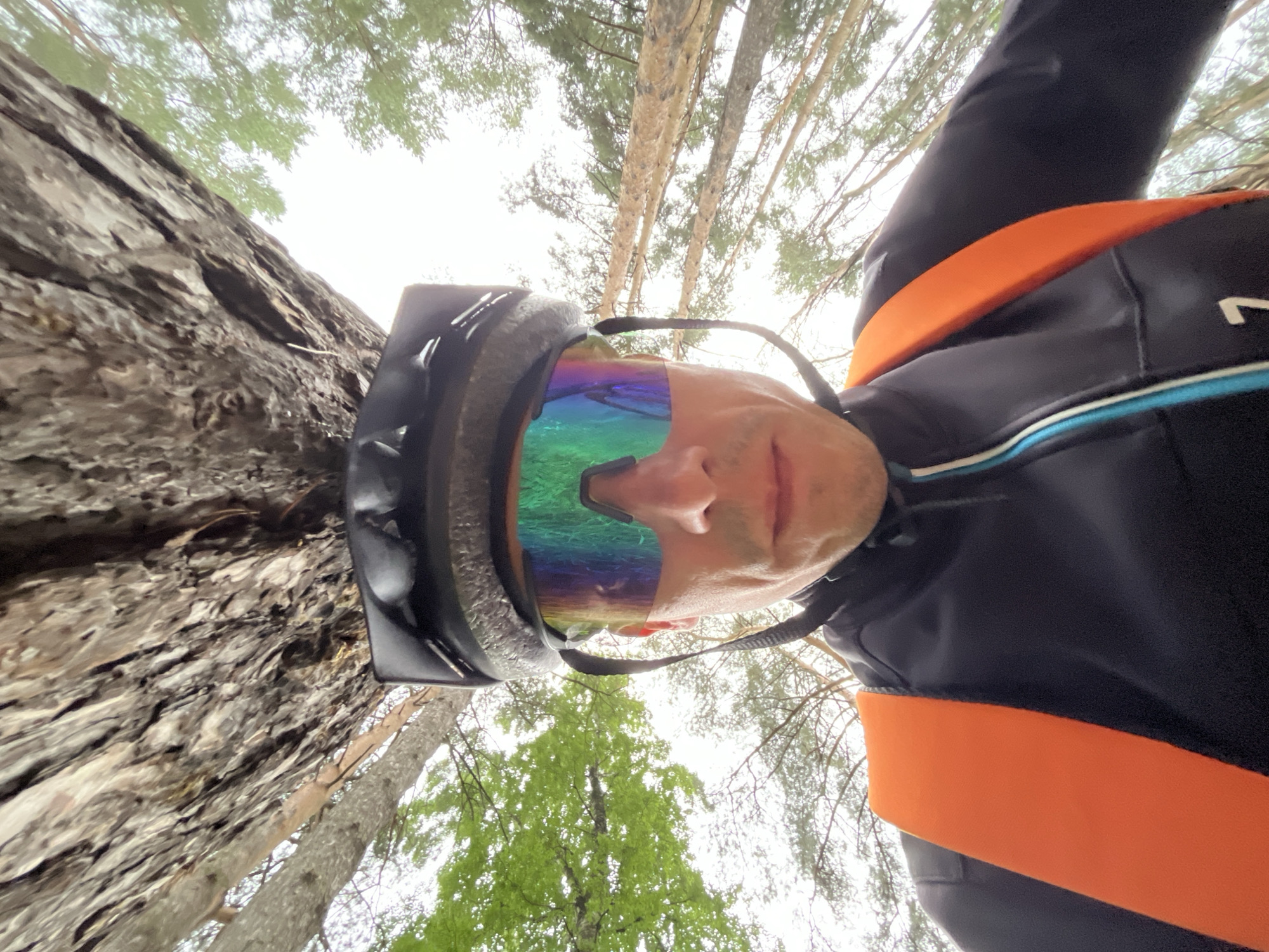 A low-angle, close-up selfie shows a person wearing a black helmet and iridescent cycling glasses. The person's face is visible, with a serious expression, and they are wearing a black cycling jacket with an orange strap across the chest. Behind the person, a large tree trunk with rough bark is prominent, and the background shows a forest canopy with green leaves and a bright, overcast sky. The image appears to be taken outdoors, likely in a natural setting. The location is identified as Chaikovsky, Russia, suggesting the scene is within a park or forest area of the city.