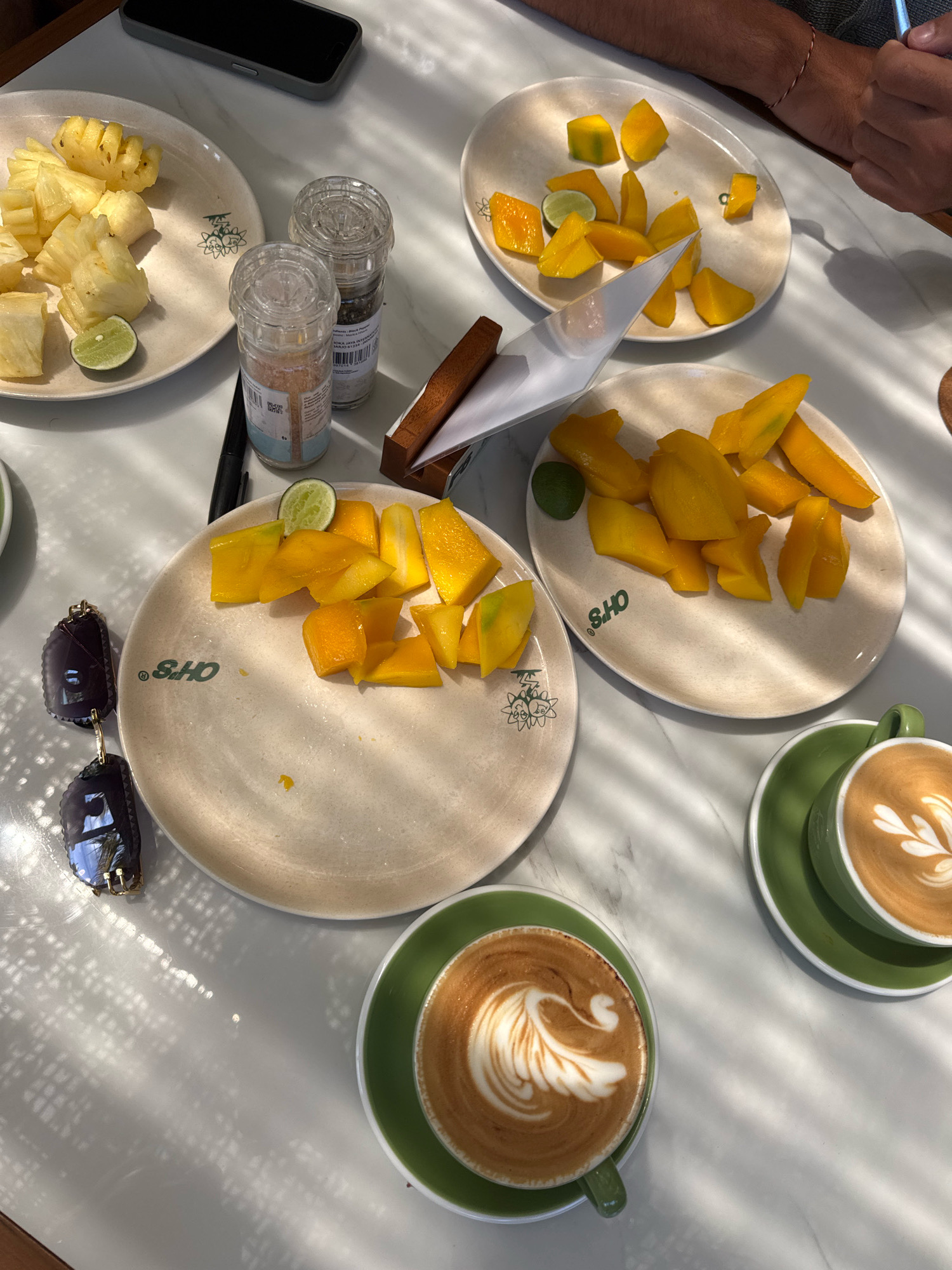 A top-down, eye-level shot captures a bright, sun-drenched table setting with an assortment of fruits and two cups of latte art coffee. The table surface is white, speckled with light and shadow patterns from an unseen source, possibly blinds or a screen.
On the table, three light-colored plates are visible. The plate in the upper left corner holds chopped pineapple pieces and two lime wedges. Adjacent to this, a pair of salt and pepper grinders stands. In the upper center, a plate is filled with sliced mangoes and a lime wedge. Another plate, in the lower center, also displays sliced mangoes and a lime wedge. To the left of this plate, a pair of dark-rimmed sunglasses rests.
In the foreground, two green ceramic cups with saucers hold cappuccinos, each adorned with intricate latte art. One coffee cup is positioned to the left, and the other to the right, slightly further back. A phone lies on the table behind the pineapple plate. A small wooden holder with a paper napkin is placed between the salt and pepper grinders and the mango plate.
The overall scene suggests a casual breakfast or snack, likely at an outdoor cafe or a well-lit indoor space. The presence of multiple plates of fruit and coffee implies a gathering of at least two people. One person's arm, with a thin red bracelet, is visible in the upper right corner, suggesting they are seated at the table.
