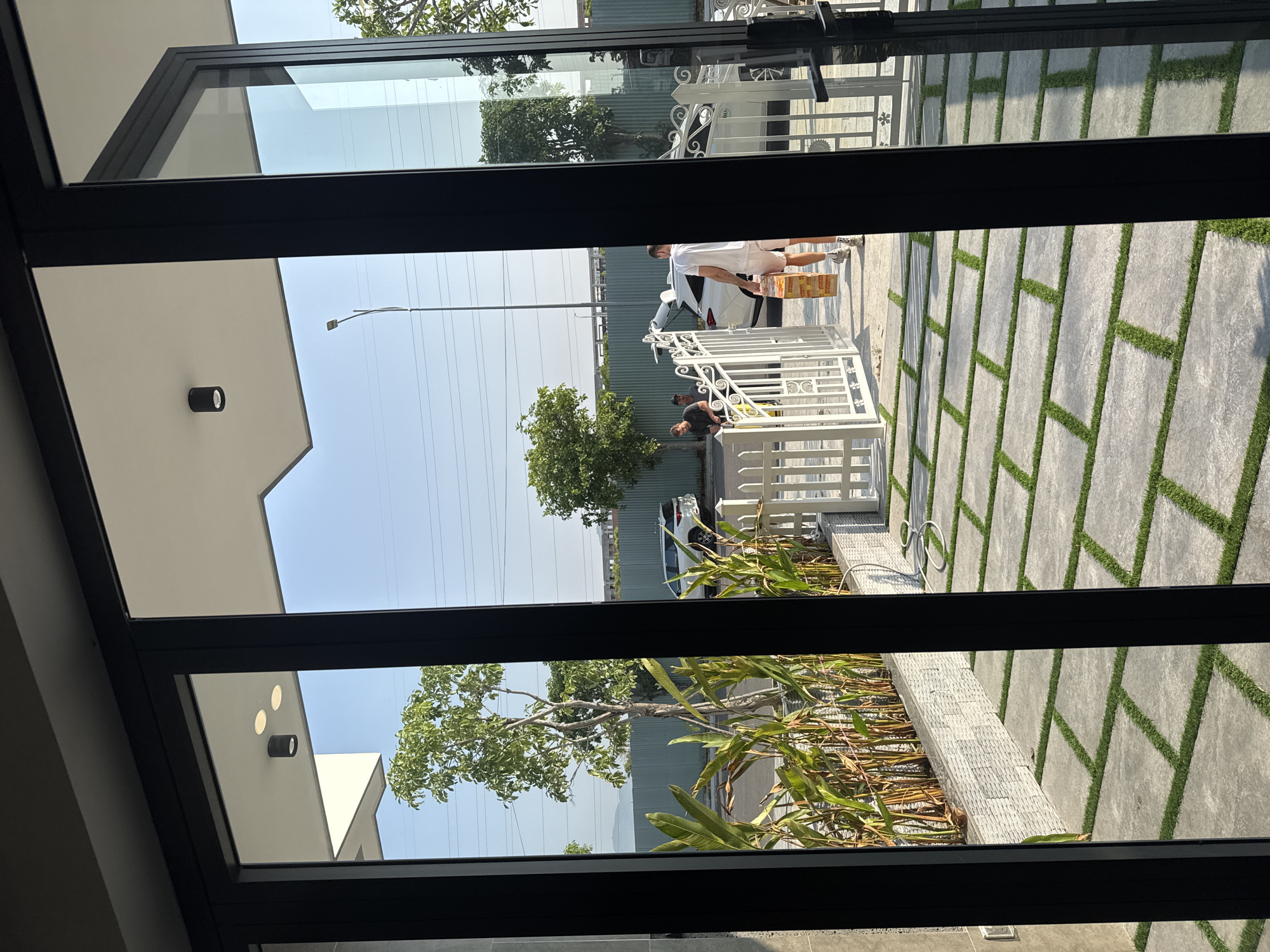 Here's a matter-of-fact description of the image:
The image is taken from indoors, looking out through a set of open black-framed glass doors or windows. The view reveals an exterior scene. Two people are visible outside. One person, dressed in white shorts and a white shirt, is carrying a box. Another person appears to be working near a white, ornate metal gate. A white car is parked nearby. A green, galvanized metal fence lines the perimeter of the property in the background. Trees are scattered around the area. The ground is covered with gray pavers separated by strips of grass. A wall covered in vegetation and rock is also present. Overhead power lines are strung in the sky. The exact location within the city of Unknown, Unknown is not discernible from the image.