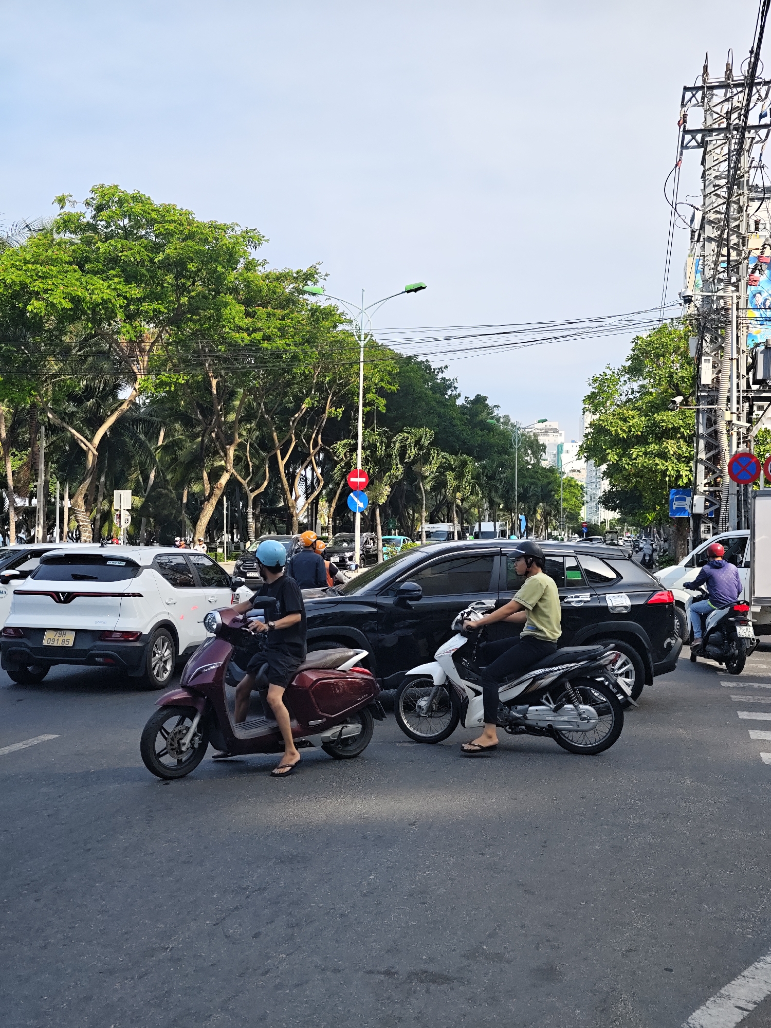 In Nha Trang Ward, Vietnam, on a paved road, a traffic scene unfolds. The sky is a light blue, with a few white clouds. Several vehicles are present, including a white SUV with license plate "79H 091.85" and a black SUV. Two individuals are riding motorcycles. One motorcycle is maroon, and the rider is wearing a black t-shirt, black shorts, and a blue helmet. The second motorcycle is white, and the rider is wearing a light green t-shirt, black pants, and a black helmet. Both riders appear to be stopped or moving slowly. Another motorcycle with a rider wearing a purple hoodie and a red helmet is visible further down the road, on the right. Traffic lights and street signs are also visible, indicating a city intersection. Lush green trees and palm trees line the sides of the road, with power lines and utility poles overhead. Buildings can be seen in the distance. The overall scene depicts a typical urban traffic environment in Vietnam.