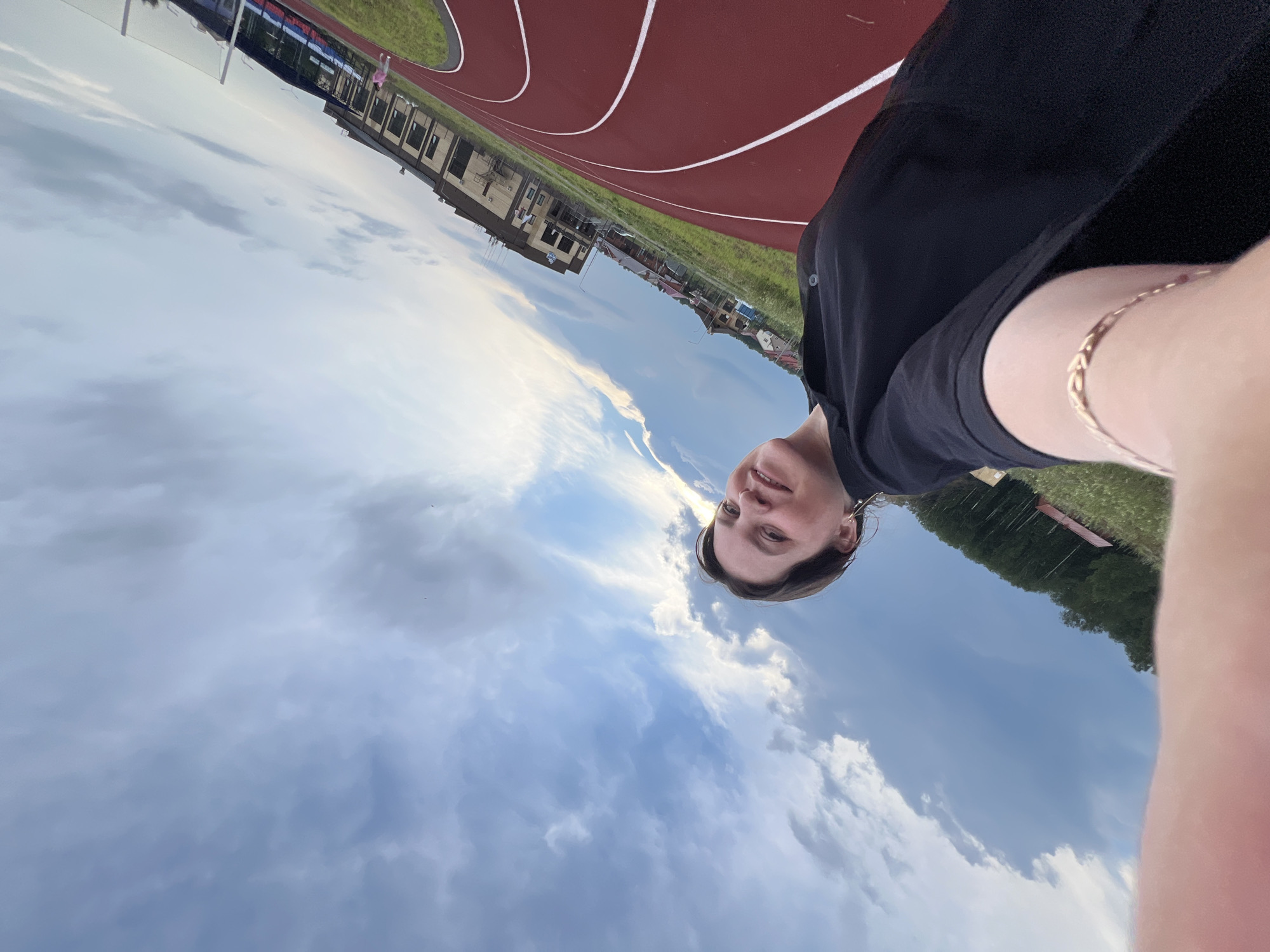 A woman with short brown hair, wearing a black shirt and a gold bracelet, smiles at the camera as she takes a selfie. She is standing on a red running track with white lines, on the grounds of a sports facility. In the background, there are buildings, green trees, and a cloudy sky with streaks of light. The image appears to be taken in Noviy, Russia, possibly at a local stadium or sports complex.
