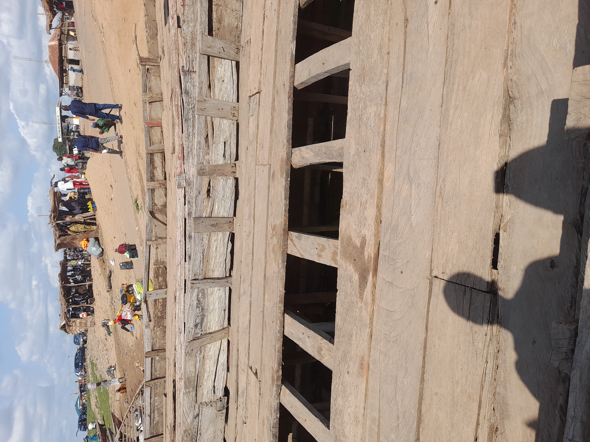 In Ibi, Nigeria, this image captures a bustling riverside scene characterized by activity and commerce. In the foreground, the weathered wooden planks of a boat are prominently featured, with visible gaps revealing the boat's interior structure. The texture of the wood, aged and worn, suggests a long history of use.
Beyond the boat, the environment transitions to a sandy embankment populated with various individuals and elements indicative of a market or transportation hub. Several makeshift stalls with thatched roofs are visible, housing vendors and customers engaged in transactions. Commodities such as fruits and bags of goods are displayed. Various vehicles, including cars and motorcycles, are parked in the background, suggesting transportation and logistics.
People are scattered throughout the scene, engaged in diverse activities: some are walking, others are carrying loads, and some appear to be interacting with vendors or fellow travelers. One person is seen on a yellow motorcycle, laden with bags. The sky above is partly cloudy, casting shadows that add depth to the scene. The overall impression is one of a lively, active community space, likely a point of transit or trade along the river.