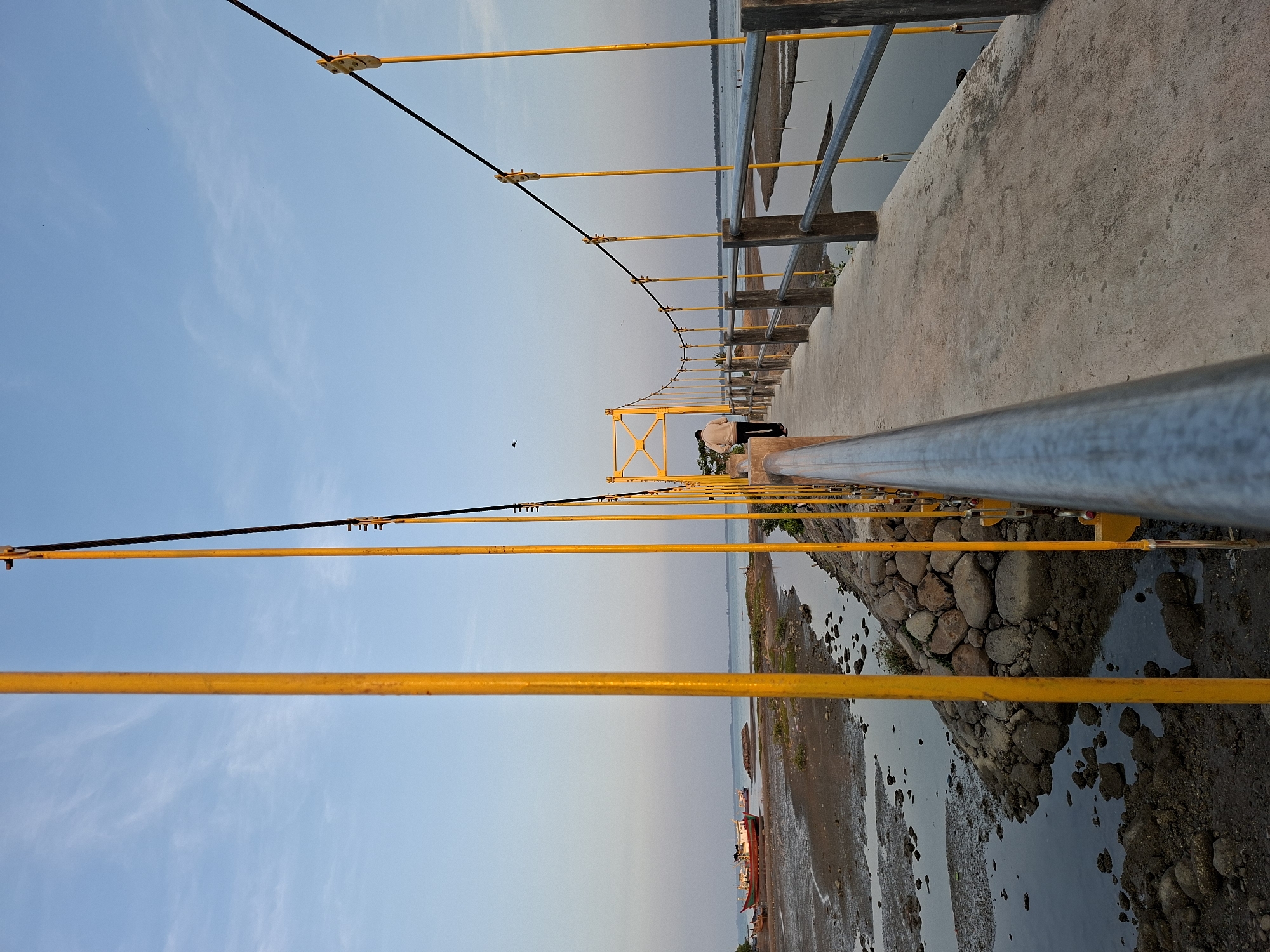 The photograph captures a long, yellow pedestrian bridge in Lhokseumawe, Indonesia, spanning a body of water. The bridge features a grey concrete path with metal railings. Vertical yellow support poles extend upward, connected by black cables. A person in light-colored clothing stands on the bridge. The water level is low, exposing mudflats and rocks along the shoreline. A couple of small boats are visible in the distance on the mudflats. The sky is light blue, suggesting either dawn or dusk.