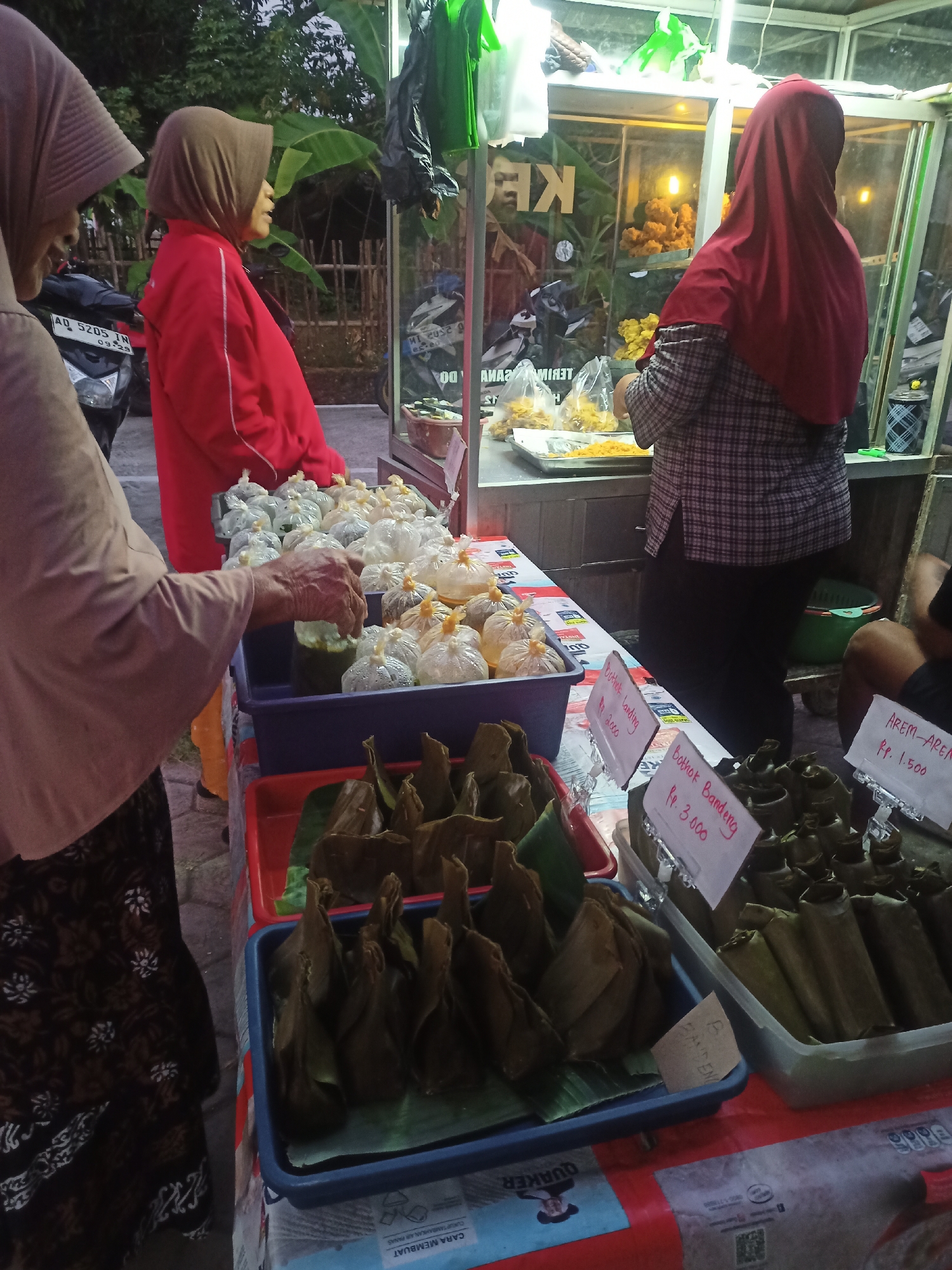 In the city of Nganti, Indonesia, at dusk, a street food vendor stall is depicted. Three women, all wearing headscarves, are present. One woman, in the foreground and on the left, is partially visible and is reaching into a green liquid container with her right hand. She is wearing a patterned dark skirt and a light brown headscarf. To her right, a second woman in a bright red jacket and brown headscarf stands, facing away from the viewer. The third woman, on the right side of the image, faces away from the camera, wearing a red headscarf and a plaid shirt. She appears to be attending to items within a glass display case that contains fried food. In front of the stall, various food items are displayed on tables. These include individually wrapped snacks in plastic bags, some appearing to be rice cakes, and other items wrapped in banana leaves. Price tags are visible for some of the food items, indicating prices such as Rp. 3.000 and Rp. 1.500. The background shows a fence, foliage, and what appears to be a parked motorcycle. The scene is lit by artificial lights from the stall.